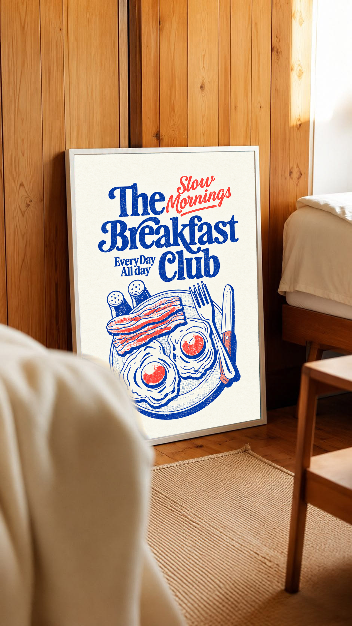 Slow Mornings Breakfast Club-Motiv mit blauer Schrift, roter Schreibschrift, zwei Spiegeleiern, Speck, Besteck und Streuern auf Creme. Im weißen Rahmen an einer holzvertäfelten Wand, neben Bett und Stuhl.