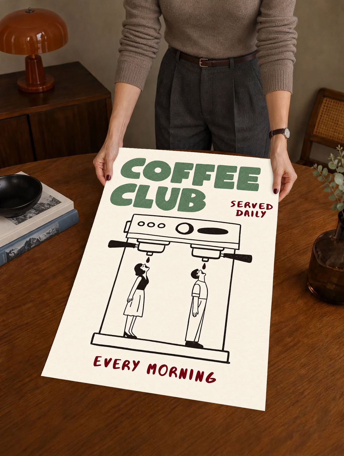 Ilustración Coffee Club verde con cafetera espresso doble negra, dos pequeñas figuras negras, palabras burdeos y fondo crema cálido. Póster sin marco sostenido sobre una mesa de madera junto a un tocadiscos.