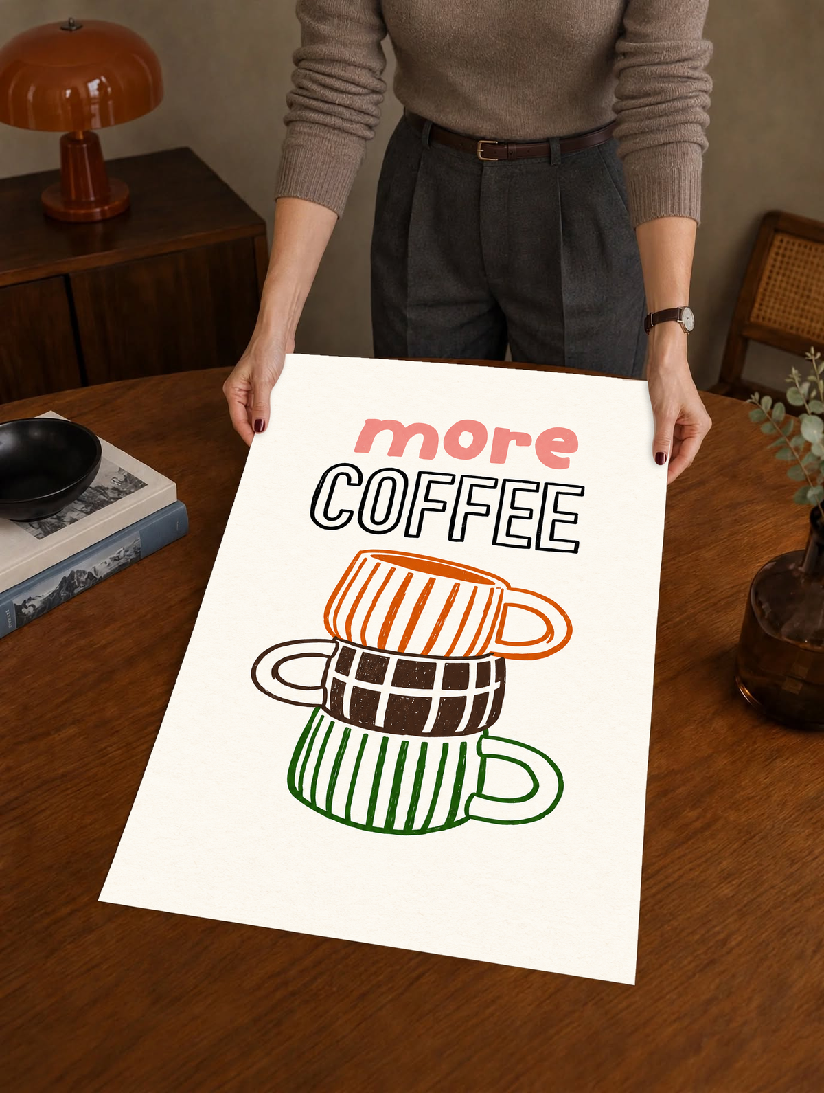 Schriftzug „More Coffee“ über drei gestapelten Tassen mit orangefarbenen Streifen, braunen Karos und grünen Streifen auf cremefarbenem Grund, als ungerahmtes Poster über einem Holztisch von einer dahinterstehenden Person gehalten.