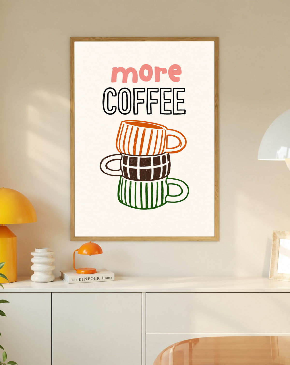 Schriftzug „More Coffee“ über drei gestapelten Tassen mit orangefarbenen Streifen, braunen Karos und grünen Streifen auf cremefarbenem Grund, in einem Eichenrahmen über einem weißen Schrank mit kleinen Lampen und Dekorationsobjekten.