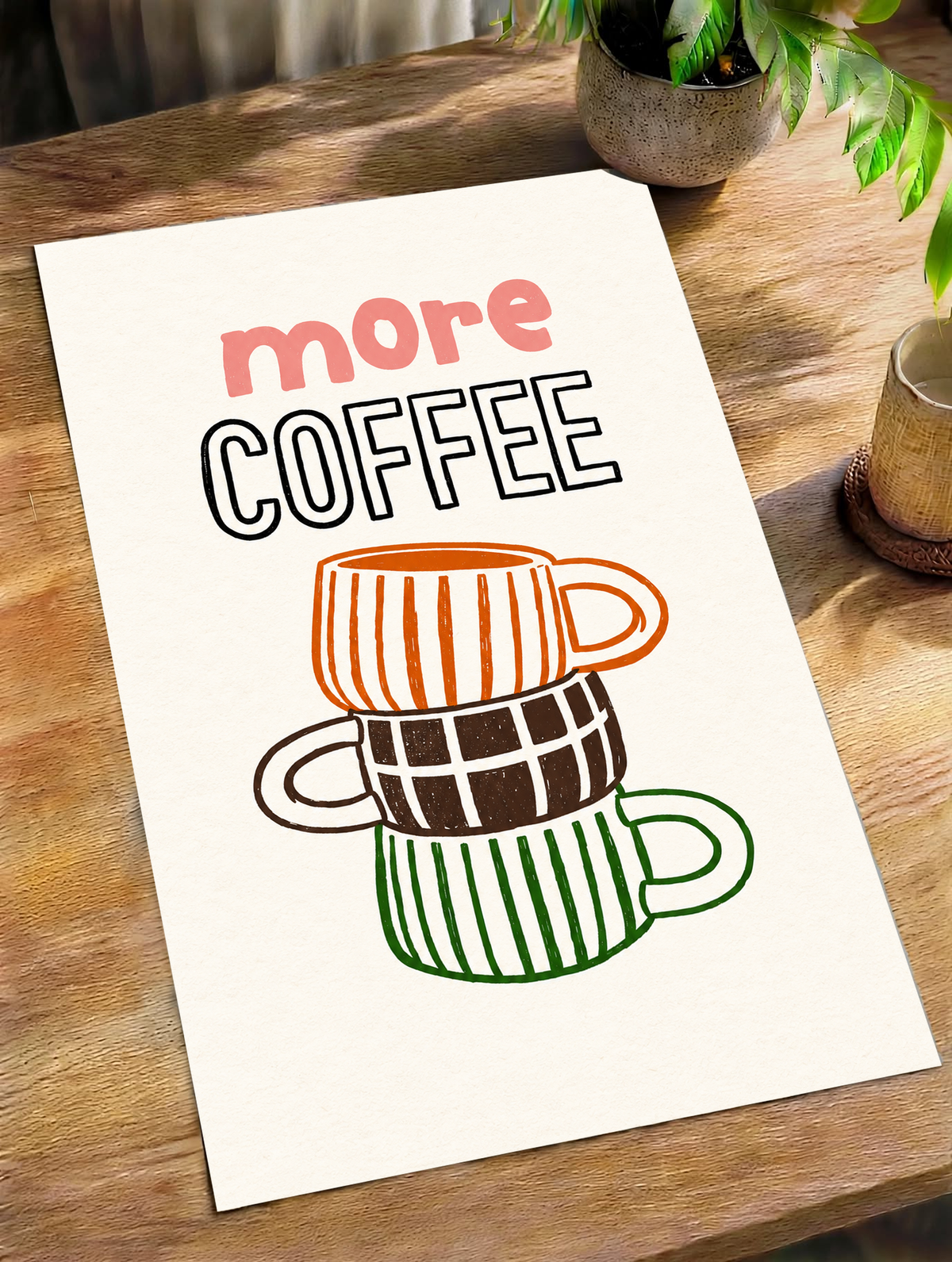 Schriftzug „More Coffee“ über drei gestapelten Tassen mit orangefarbenen Streifen, braunen Karos und grünen Streifen auf cremefarbenem Grund, als loses Poster schräg auf einem Holztisch neben Topfpflanzen liegend.