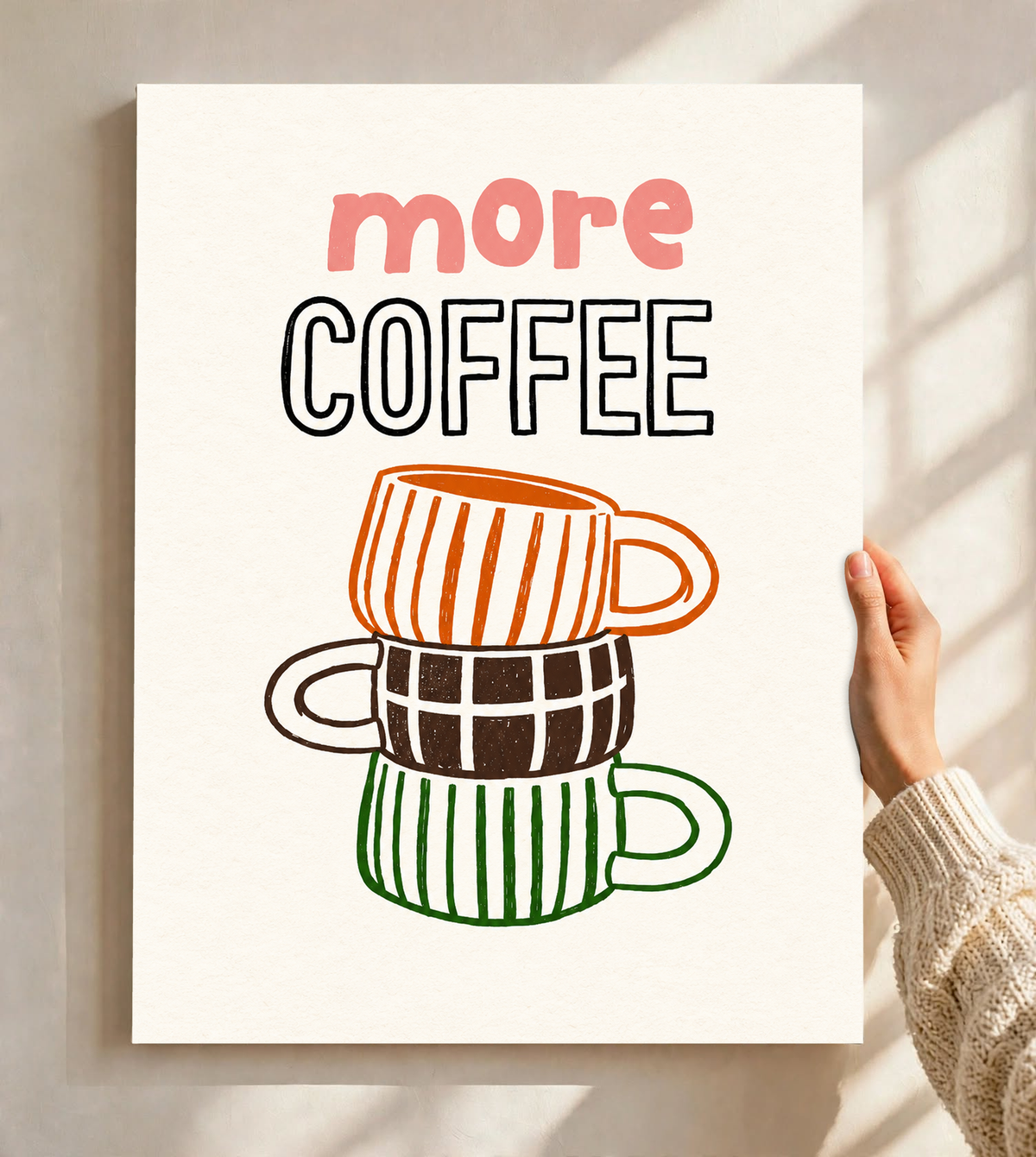 Schriftzug „More Coffee“ über drei gestapelten Tassen mit orangefarbenen Streifen, braunen Karos und grünen Streifen auf cremefarbenem Grund, als ungerahmtes Poster vor einer hellen Wand mit weichen Fensterschatten gehalten.