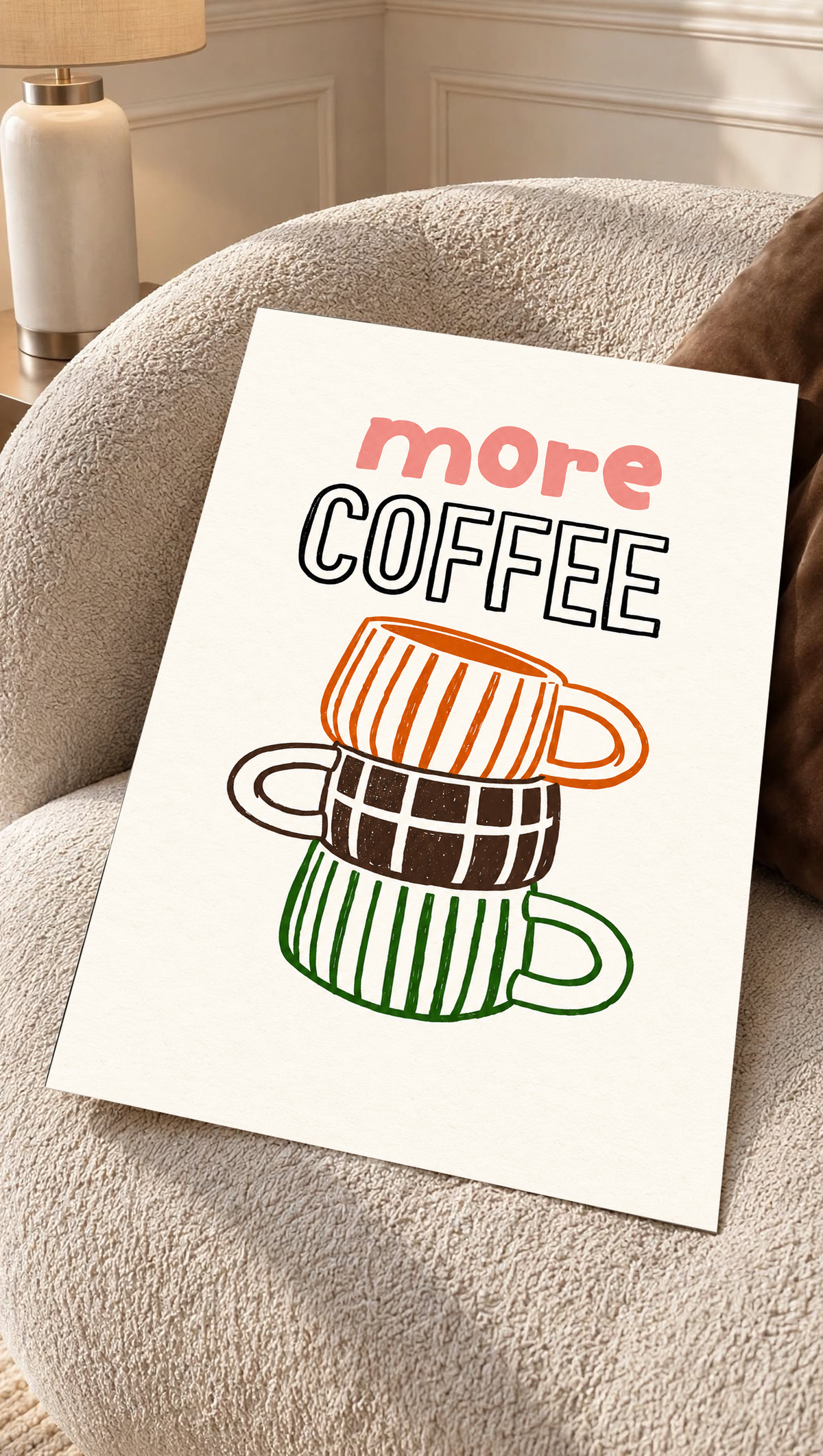 Schriftzug „More Coffee“ über drei gestapelten Tassen mit orangefarbenen Streifen, braunen Karos und grünen Streifen auf cremefarbenem Grund, als loses Poster auf einem cremefarbenen Sessel mit strukturierter Oberfläche.