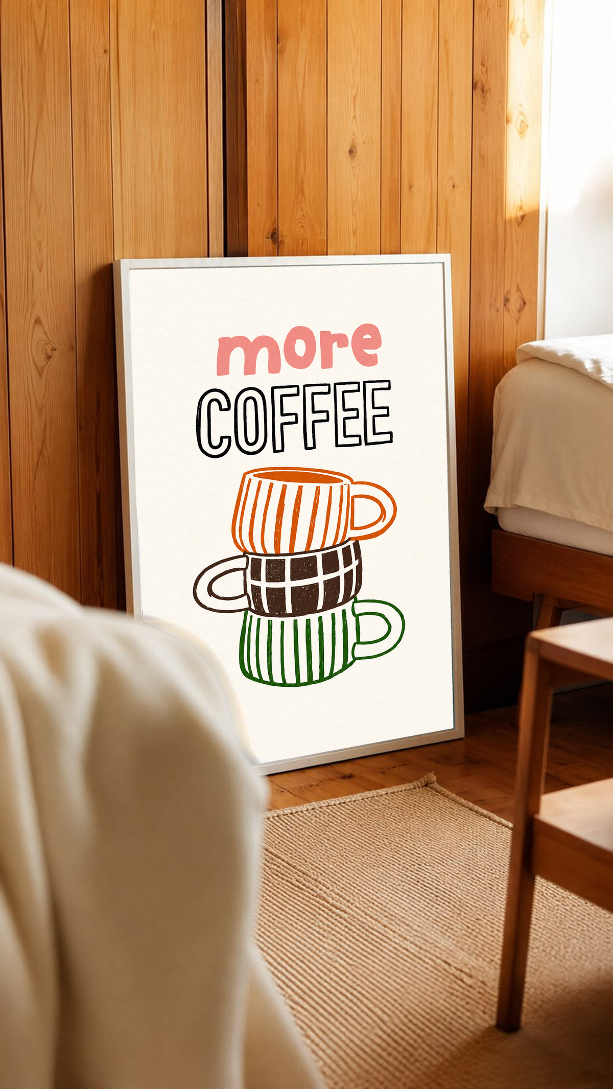 Schriftzug „More Coffee“ über drei gestapelten Tassen mit orangefarbenen Streifen, braunen Karos und grünen Streifen auf cremefarbenem Grund, in einem weißen Rahmen an einer holzverkleideten Wand neben einem Stuhl in einem sonnigen Zimmer.