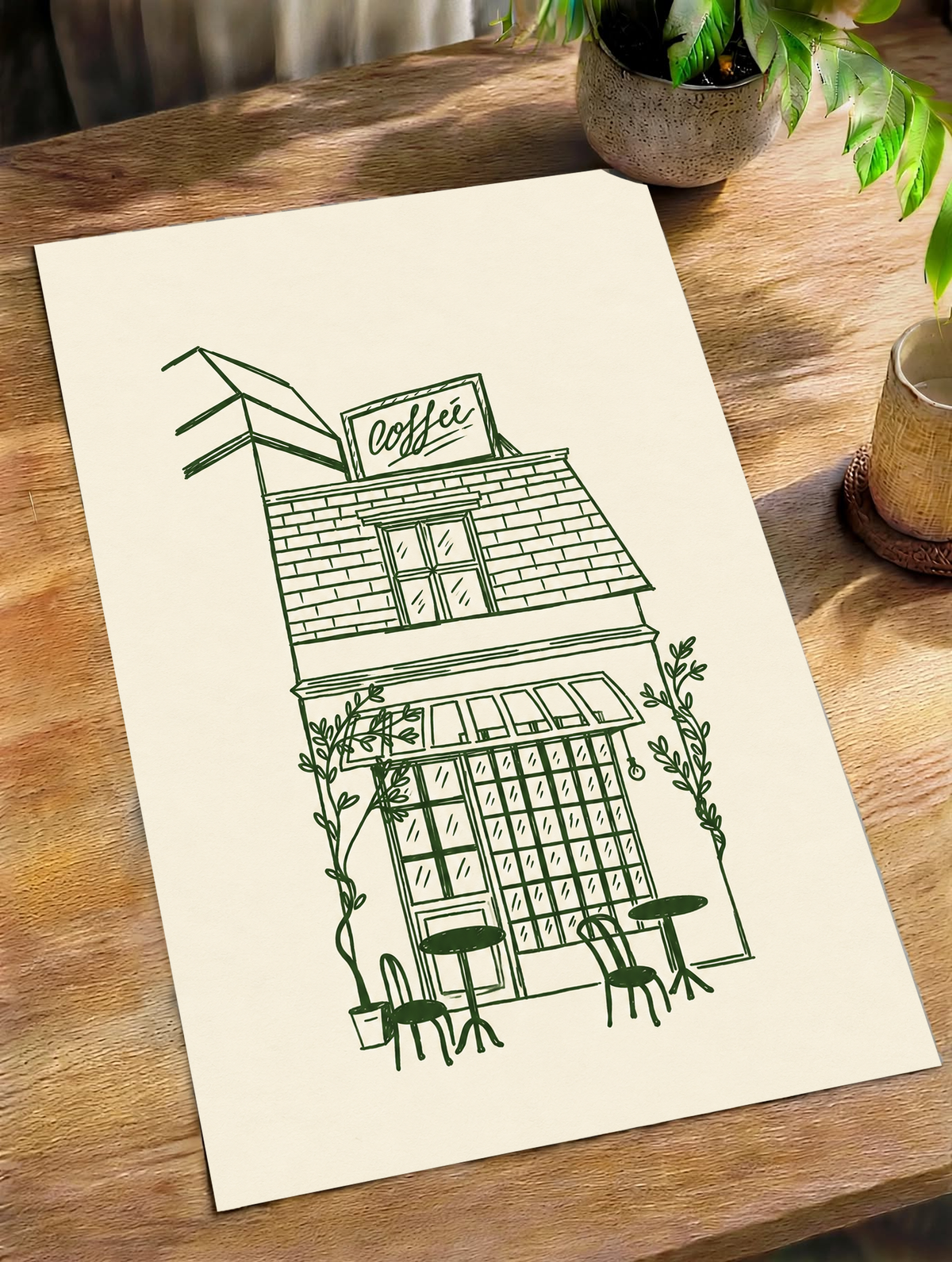 Kleines Kaffeehaus in dunkelgrünen Linien auf Creme, mit Dachschild, gestreifter Markise, Kletterpflanzen sowie Tischen und Stühlen im Freien, als loses Poster schräg auf einem Holztisch neben Topfpflanzen liegend.