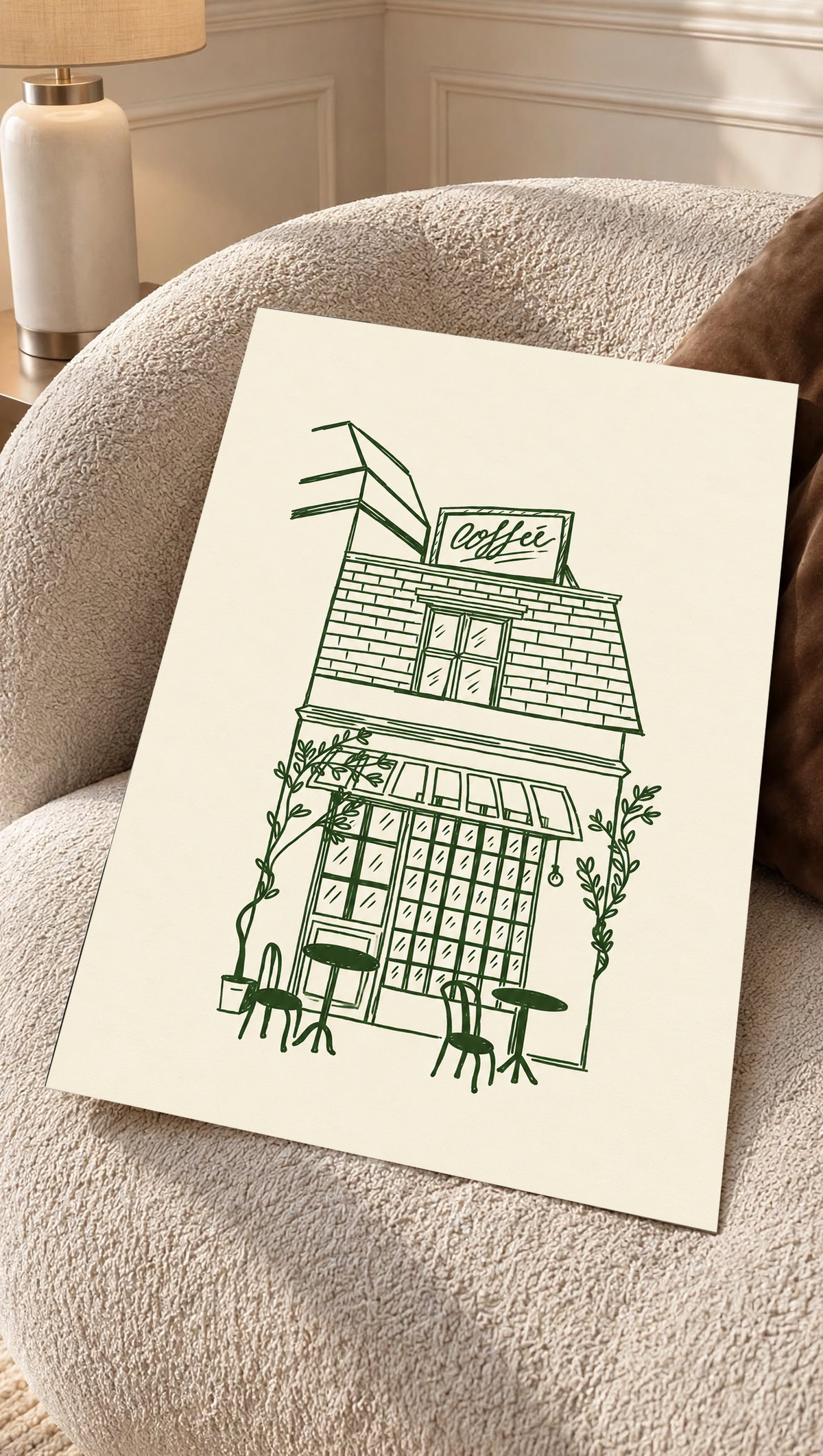 Kleines Kaffeehaus in dunkelgrünen Linien auf Creme, mit Dachschild, gestreifter Markise, Kletterpflanzen sowie Tischen und Stühlen im Freien, als loses Poster auf einem cremefarbenen Sessel mit strukturierter Oberfläche.