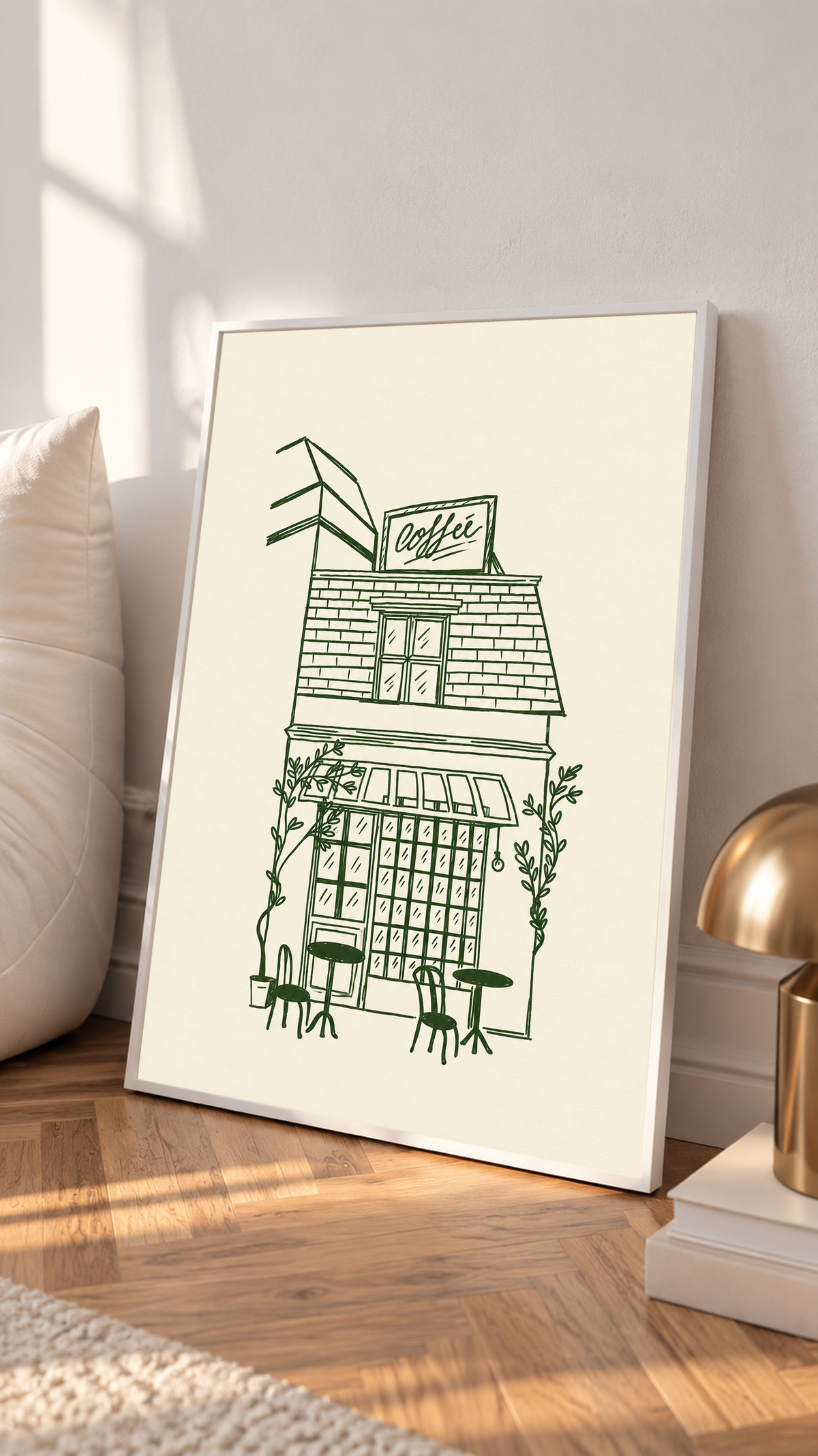 Kleines Kaffeehaus in dunkelgrünen Linien auf Creme, mit Dachschild, gestreifter Markise, Kletterpflanzen sowie Tischen und Stühlen im Freien, in einem weißen Rahmen auf einem Holzboden, neben einem hellen Kissen und einer messingfarbenen Lampe.