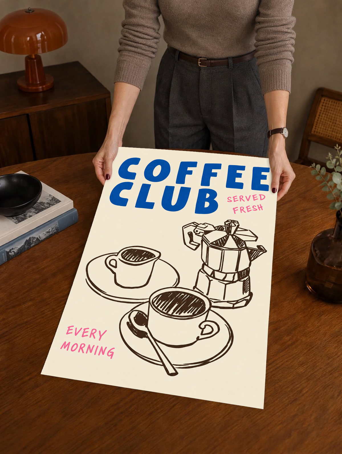 Blauer Schriftzug „Coffee Club“ mit rosa „Served Fresh“ und „Every Morning“ über einer braunen Mokkakanne und zwei Tassen auf Creme, als ungerahmtes Poster über einem Holztisch von einer dahinterstehenden Person gehalten.
