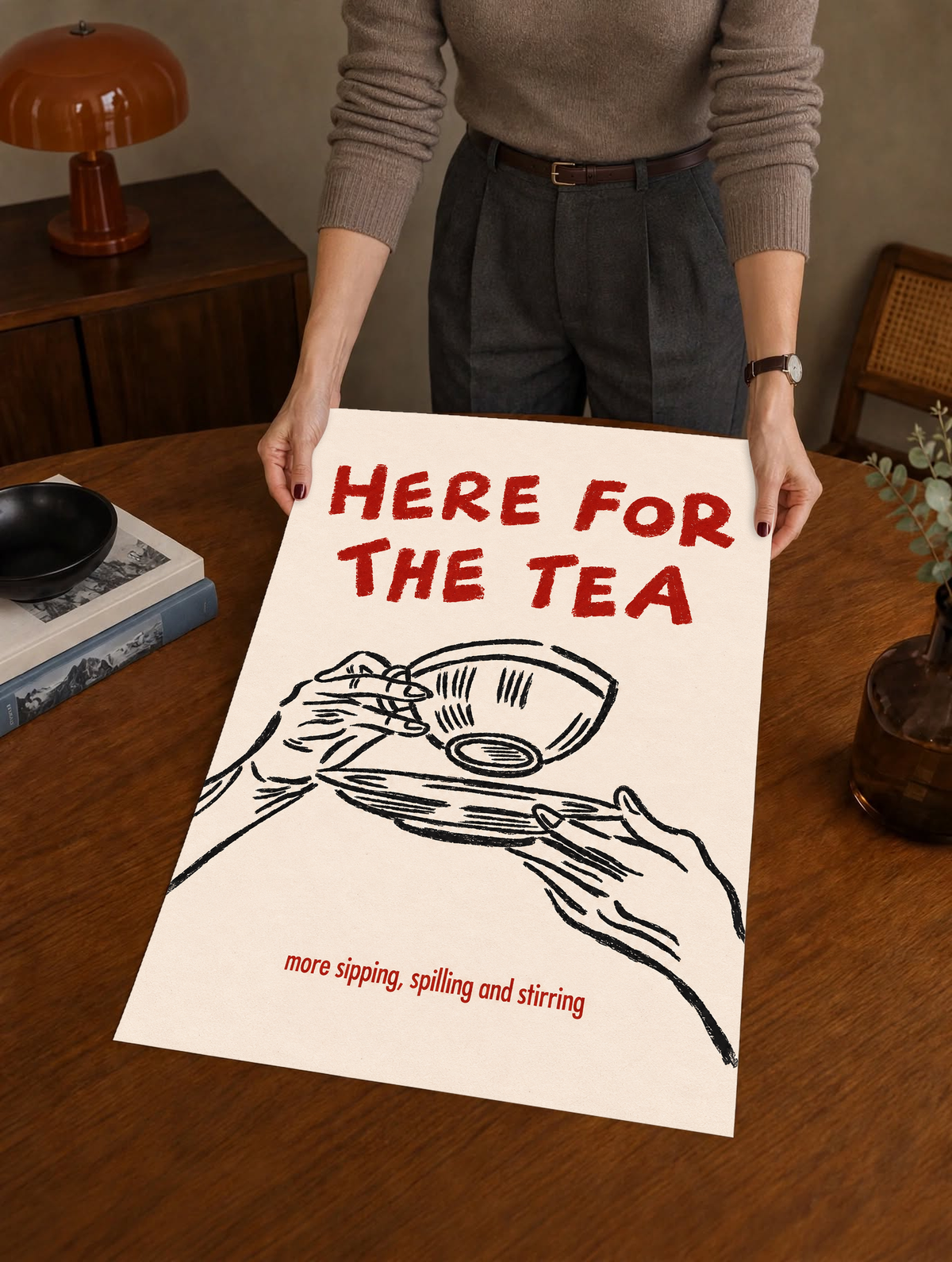 Roter Schriftzug „Here for the Tea“ über schwarz gezeichneten Händen mit Teetasse und Untertasse; darunter „more sipping, spilling and stirring“ auf Creme, als ungerahmtes Poster über einem Holztisch von einer dahinterstehenden Person gehalten.