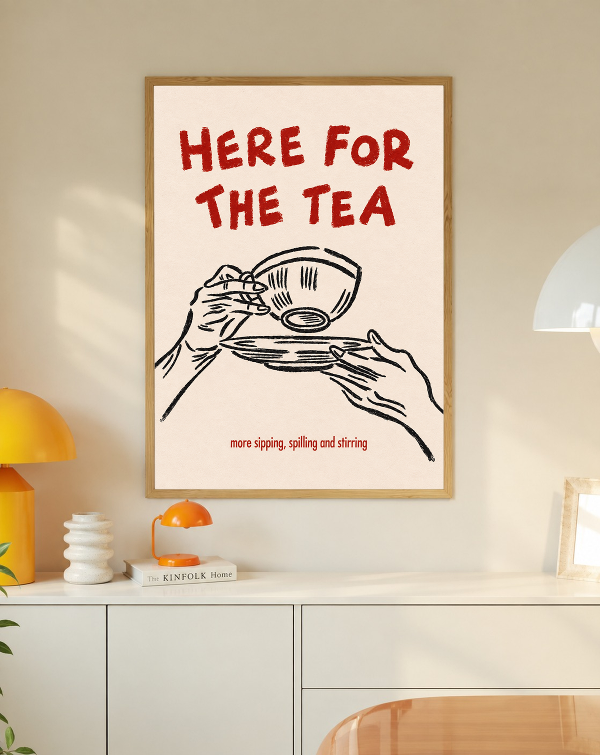 Roter Schriftzug „Here for the Tea“ über schwarz gezeichneten Händen mit Teetasse und Untertasse; darunter „more sipping, spilling and stirring“ auf Creme, in einem Eichenrahmen über einem weißen Schrank mit kleinen Lampen und Dekorationsobjekten.