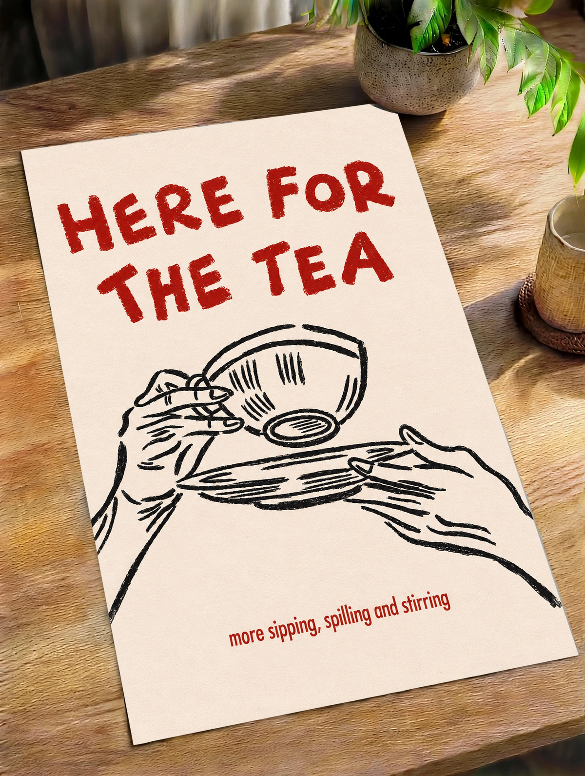 Roter Schriftzug „Here for the Tea“ über schwarz gezeichneten Händen mit Teetasse und Untertasse; darunter „more sipping, spilling and stirring“ auf Creme, als loses Poster schräg auf einem Holztisch neben Topfpflanzen liegend.
