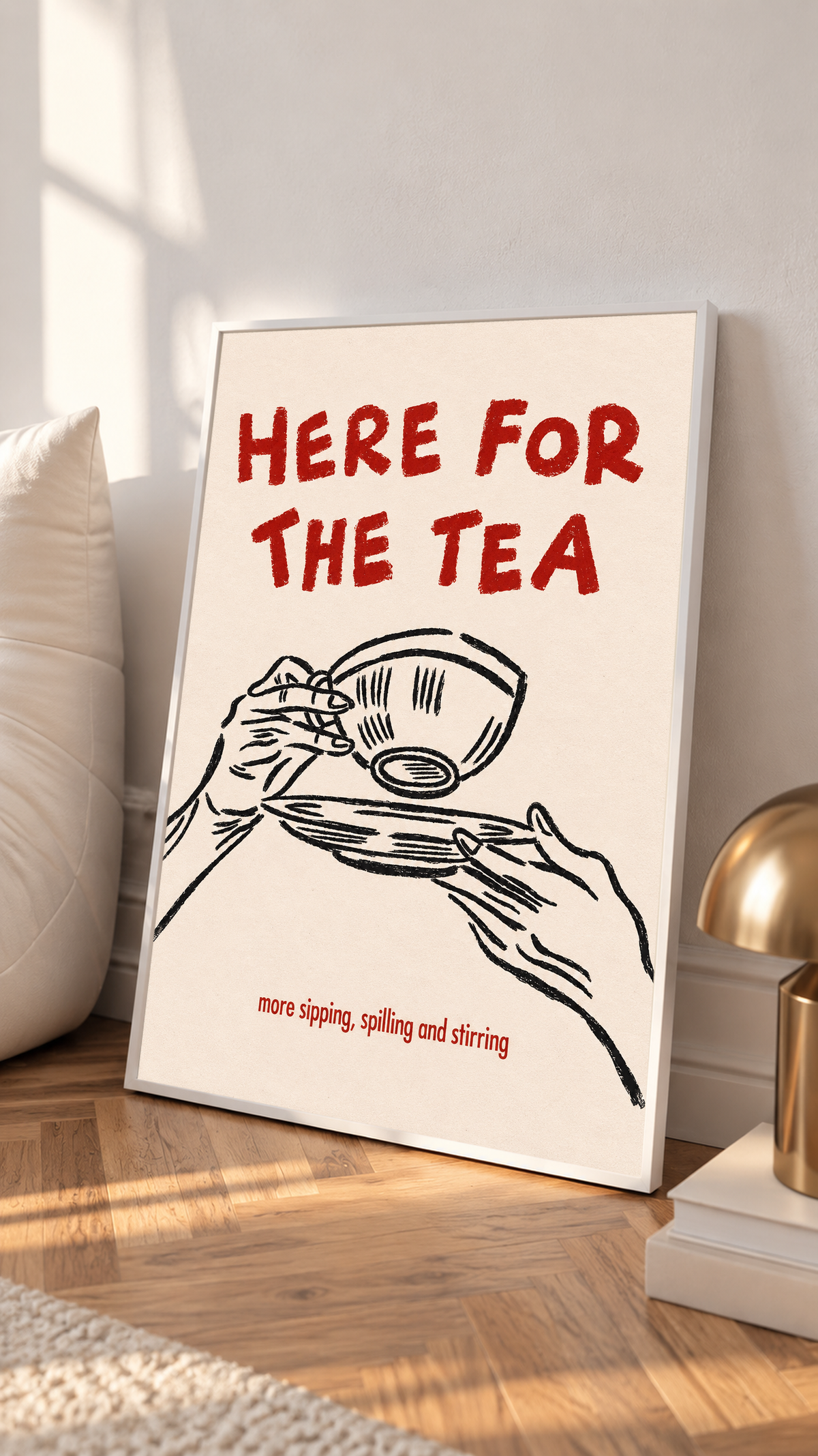 Roter Schriftzug „Here for the Tea“ über schwarz gezeichneten Händen mit Teetasse und Untertasse; darunter „more sipping, spilling and stirring“ auf Creme, in einem weißen Rahmen auf einem Holzboden, neben einem hellen Kissen und einer messingfarbenen Lampe.