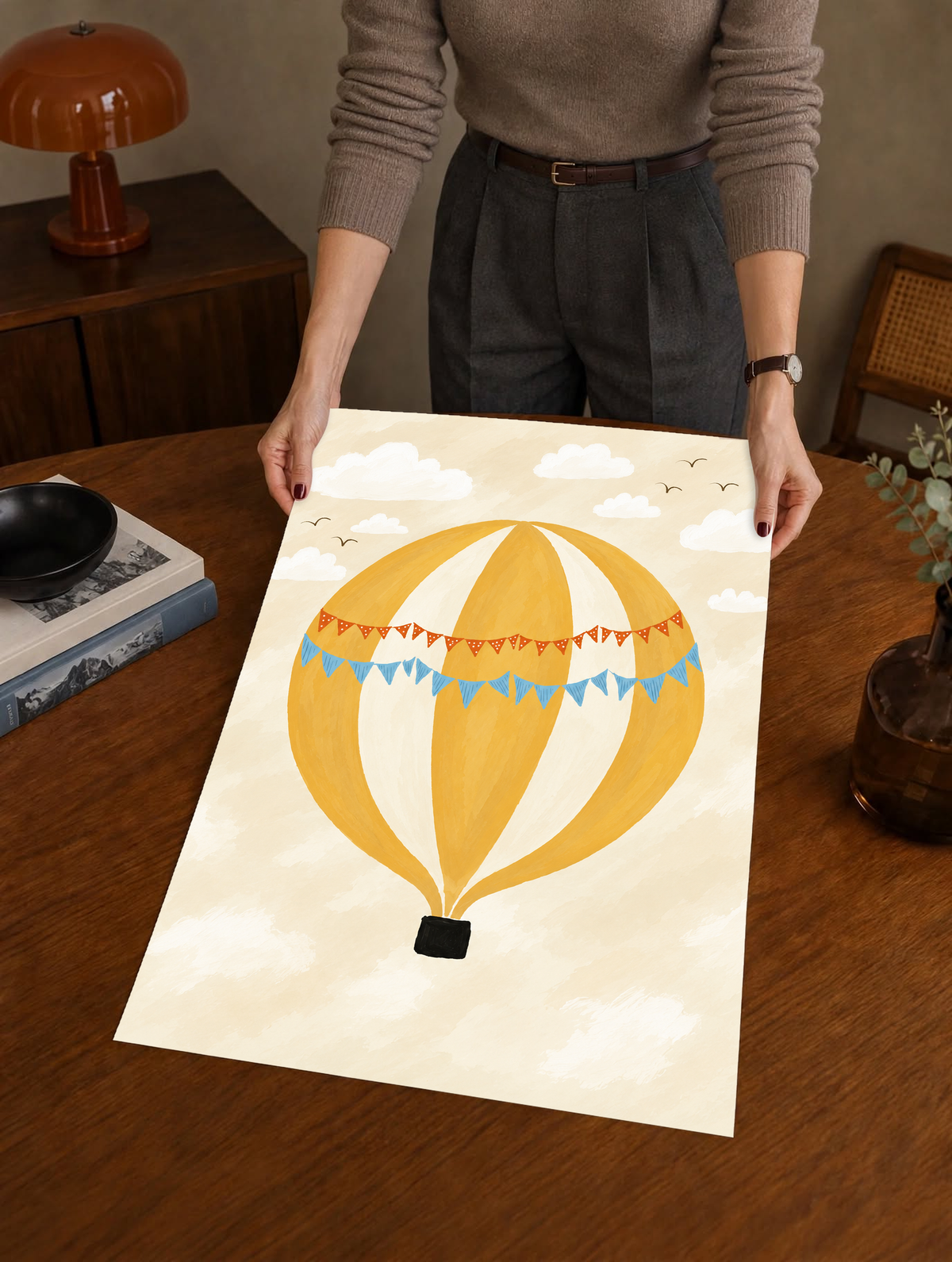 Globo aerostático amarillo y crema con banderines azules y otros naranjas con puntos blancos, cesta negra, nubes blancas y pájaros en un cielo crema. Como póster sin marco sostenido sobre una mesa de madera, con una lámpara marrón detrás.