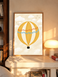 Globo aerostático amarillo y crema con banderines azules y otros naranjas con puntos blancos, cesta negra, nubes blancas y pájaros en un cielo crema. En un marco de tono roble sobre una repisa junto a una lámpara encendida.