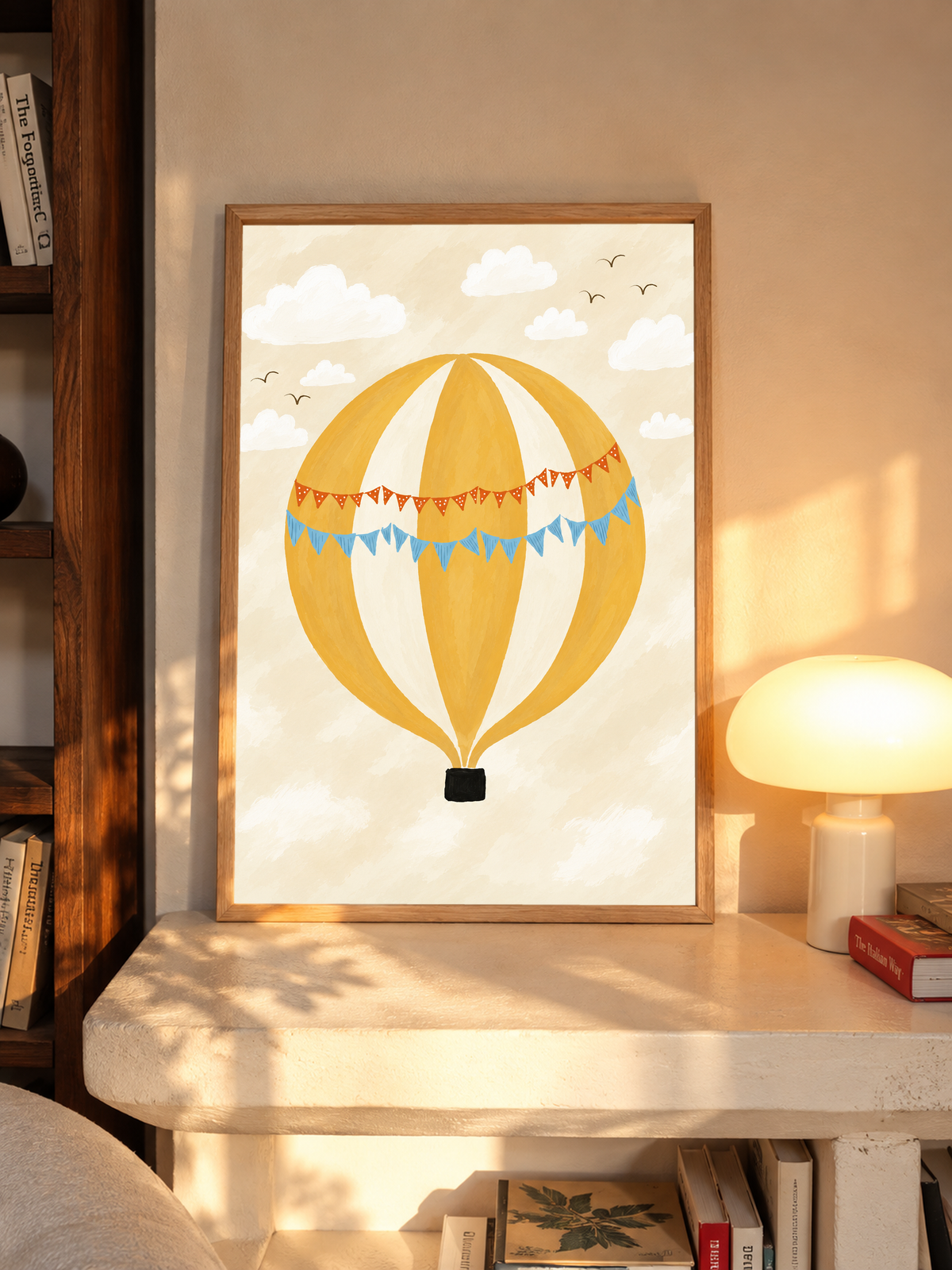 Globo aerostático amarillo y crema con banderines azules y otros naranjas con puntos blancos, cesta negra, nubes blancas y pájaros en un cielo crema. En un marco de tono roble sobre una repisa junto a una lámpara encendida.