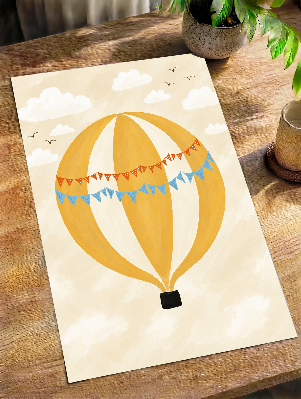 Globo aerostático amarillo y crema con banderines azules y otros naranjas con puntos blancos, cesta negra, nubes blancas y pájaros en un cielo crema. Póster sin marco sobre una mesa de madera junto a una planta de hojas verdes y una taza.