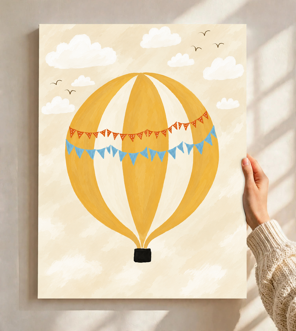 Globo aerostático amarillo y crema con banderines azules y otros naranjas con puntos blancos, cesta negra, nubes blancas y pájaros en un cielo crema. Lámina sin marco sostenida ante una pared clara con luz solar y sombras de ventana.