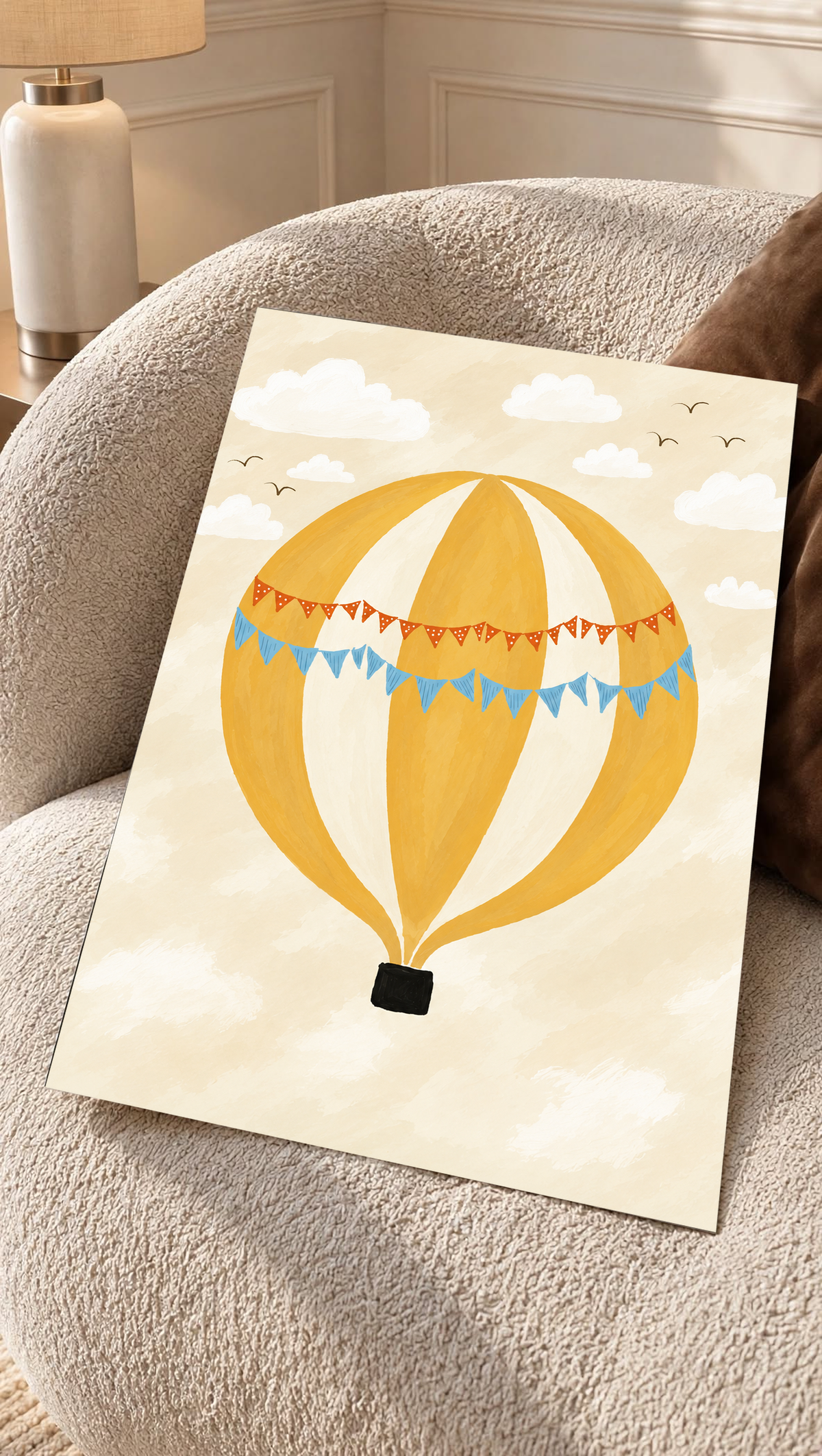 Globo aerostático amarillo y crema con banderines azules y otros naranjas con puntos blancos, cesta negra, nubes blancas y pájaros en un cielo crema. Lámina sin marco apoyada sobre un sillón de textura color crema.