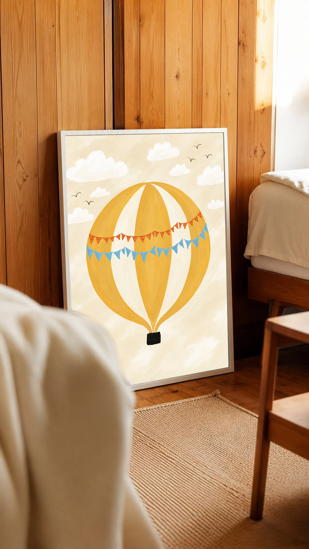 Globo aerostático amarillo y crema con banderines azules y otros naranjas con puntos blancos, cesta negra, nubes blancas y pájaros en un cielo crema. Marco blanco junto a una cama, ante un revestimiento de madera vertical.