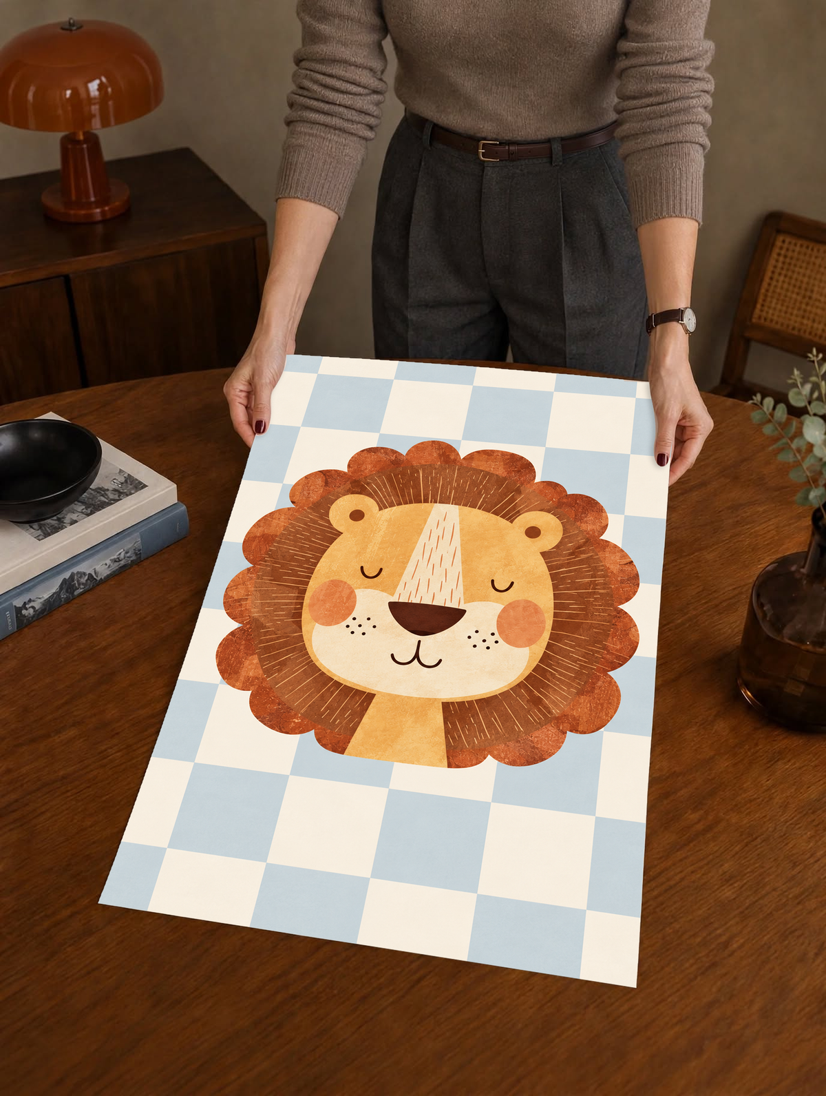 Rostro de león dorado con ojos cerrados, mejillas naranjas y melena ondulada marrón óxido sobre un damero azul claro y crema. Como póster sin marco sostenido sobre una mesa de madera, con una lámpara marrón detrás.