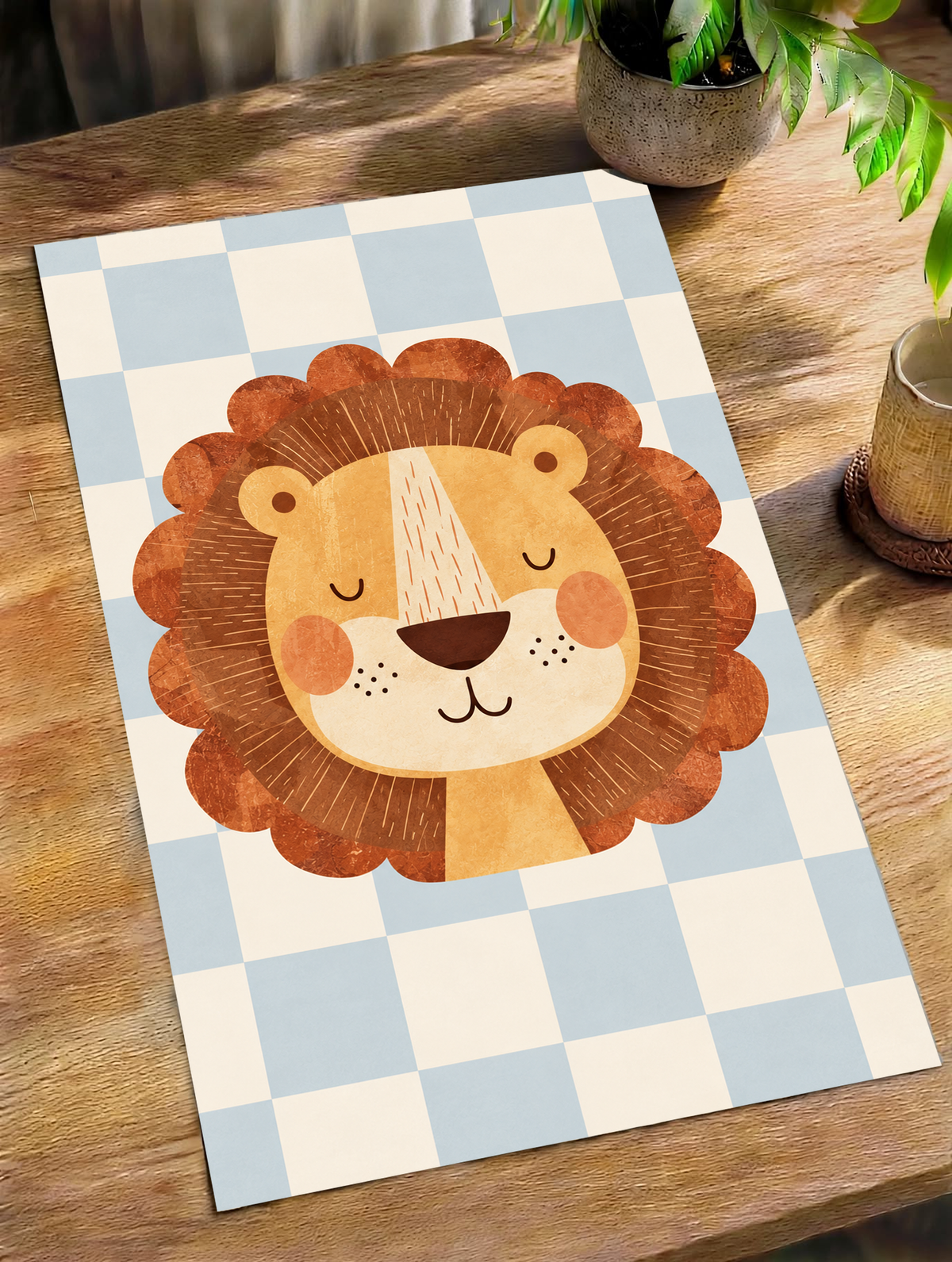Rostro de león dorado con ojos cerrados, mejillas naranjas y melena ondulada marrón óxido sobre un damero azul claro y crema. Póster sin marco sobre una mesa de madera junto a una planta de hojas verdes y una taza.