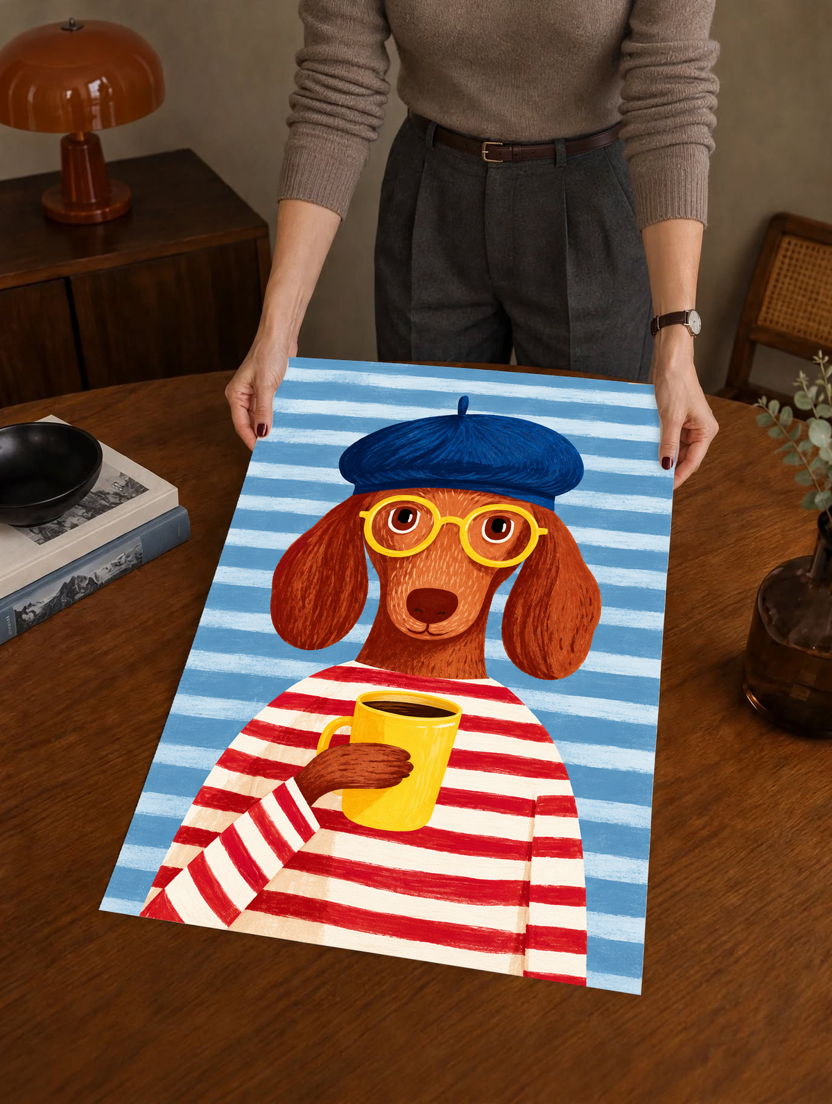 Póster sin marco sostenido por una persona sobre una mesa de madera: Un perro marrón de orejas largas, boina azul, gafas amarillas y camiseta de rayas rojas y crema sostiene una taza amarilla ante un fondo de rayas azules.