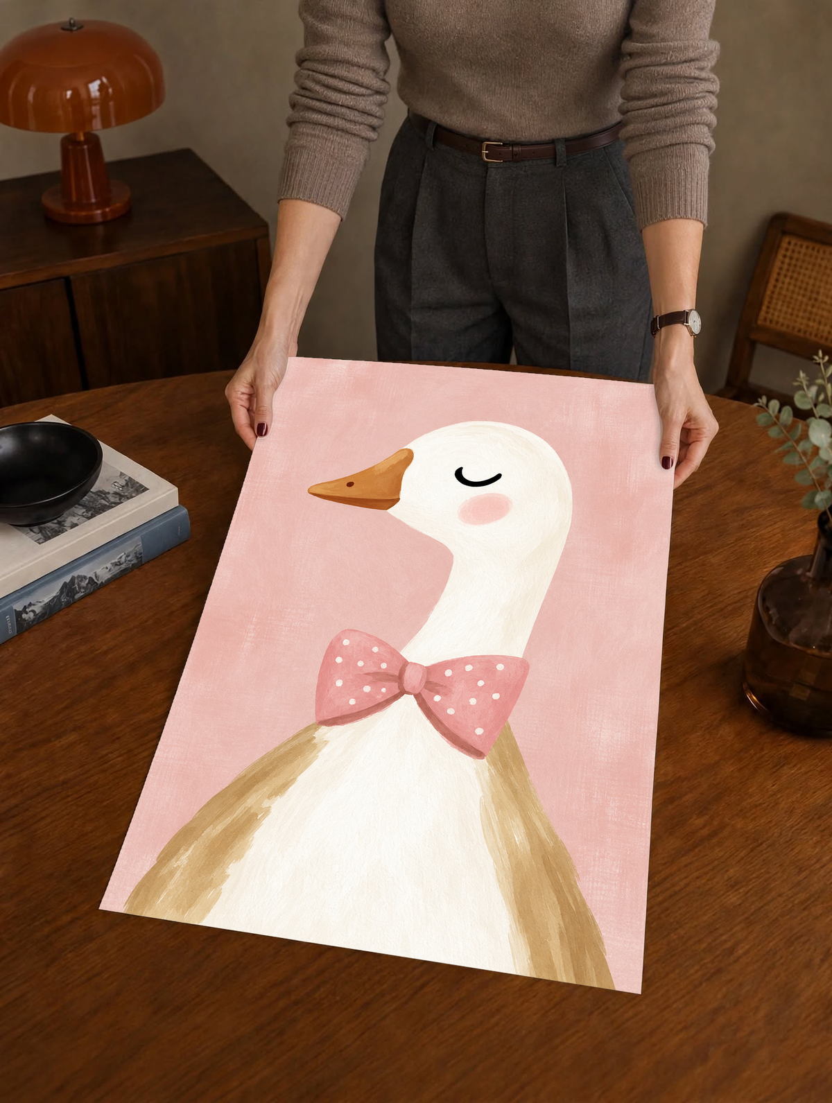 Primer plano de oca blanca con ojo cerrado, mejilla rosada, pico ocre y lazo rosa de lunares blancos sobre fondo rosa suave. Como póster sin marco sostenido sobre una mesa de madera, con una lámpara marrón detrás.