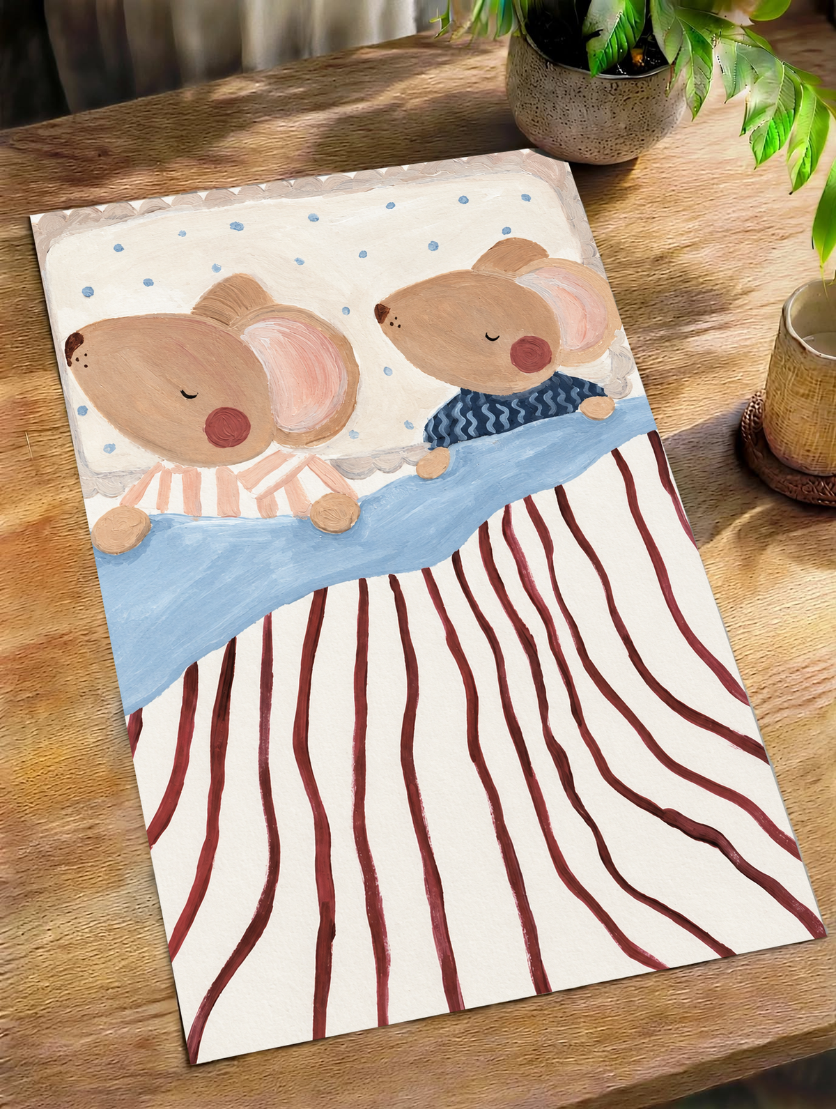 Dos ratones marrones dormidos y de mejillas rosadas, en pijamas de rayas melocotón y ondas azul marino, con ropa de cama azul, rayas burdeos y almohada punteada. Póster sin marco sobre una mesa de madera junto a una planta de hojas verdes y una taza.