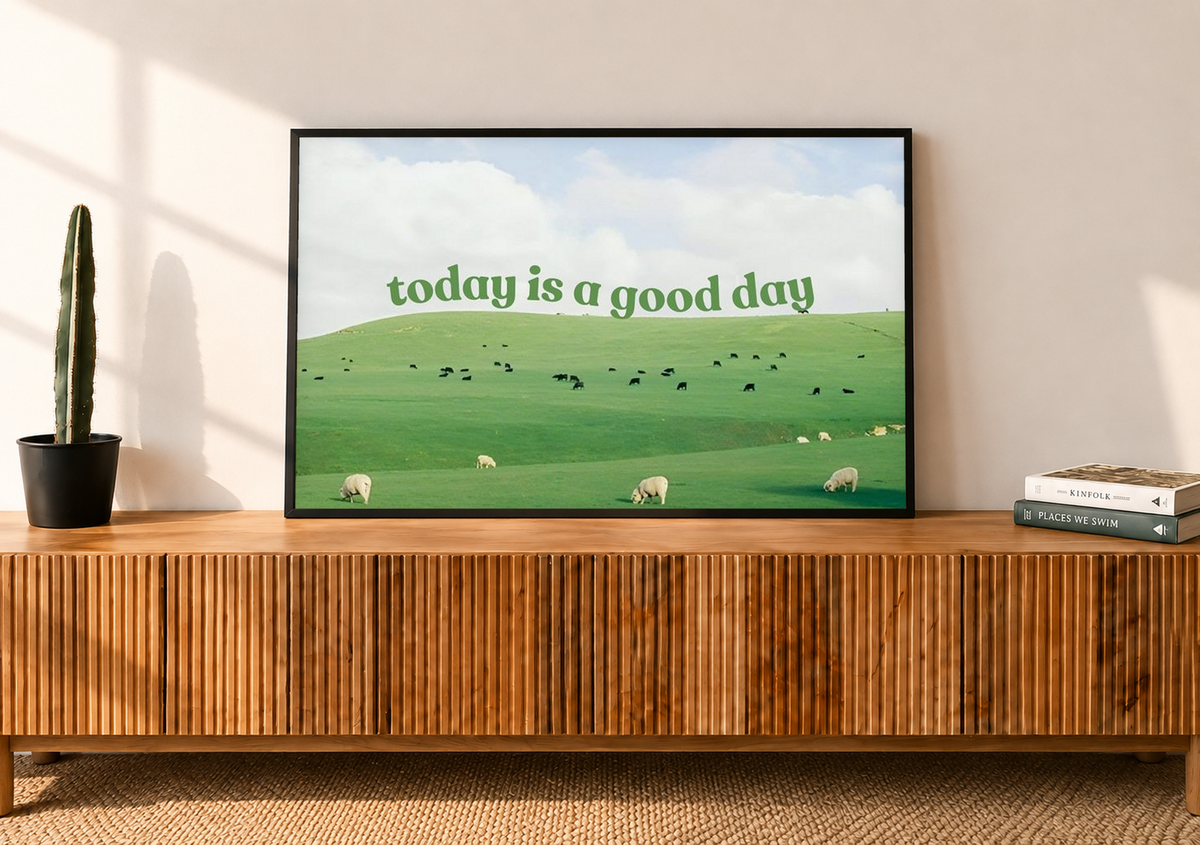 Grüne Schrift 'today is a good day' über Weiden mit Schafen, kleinen Rindern und hellem Wolkenhimmel. Schwarzer Rahmen auf einer Holzkommode mit Lamellenfront neben einem Kaktus im Topf und Büchern.