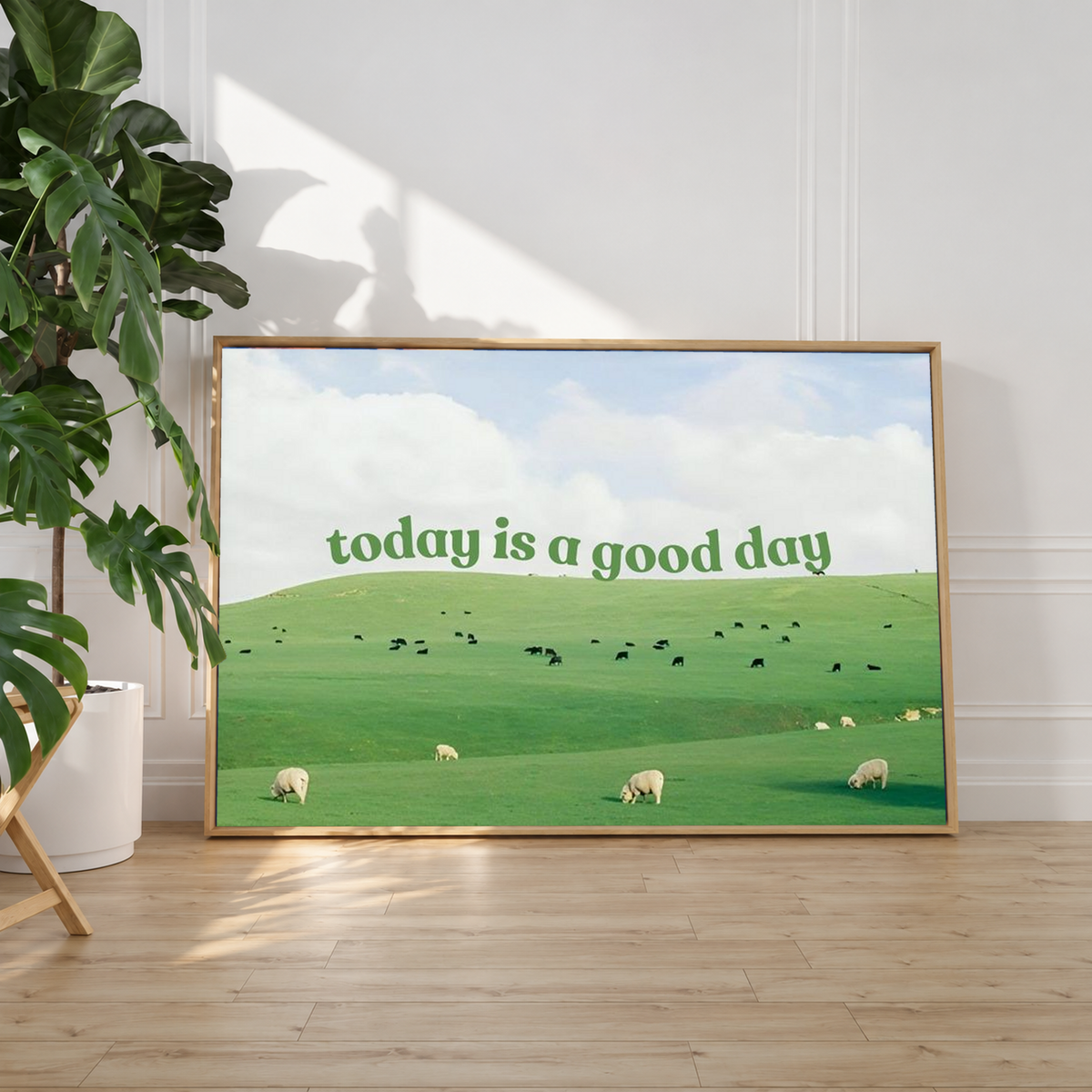 Grüne Schrift 'today is a good day' über Weiden mit Schafen, kleinen Rindern und hellem Wolkenhimmel. Heller Schattenfugenrahmen an einer weißen Wand neben einer Zimmerpflanze; Sonnenlicht auf dem Holzboden.