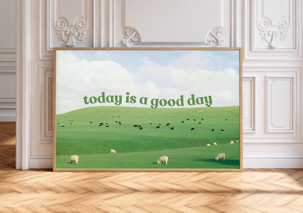 Grüne Schrift 'today is a good day' über Weiden mit Schafen, kleinen Rindern und hellem Wolkenhimmel. Eichenfarbener Rahmen an einer weißen Wand mit Zierleisten über einem Fischgrätboden.
