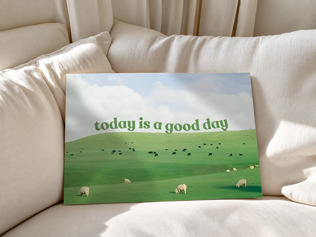 Grüne Schrift 'today is a good day' über Weiden mit Schafen, kleinen Rindern und hellem Wolkenhimmel. Ungerahmtes Poster an weiche weiße Sofakissen gelehnt.
