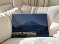 Warm beleuchteter Laden mit blauer Fassade unter einem verschneiten Berg im Abendblau, mit Person, Auto und Schnee. Ungerahmtes Poster an weiche weiße Sofakissen gelehnt.