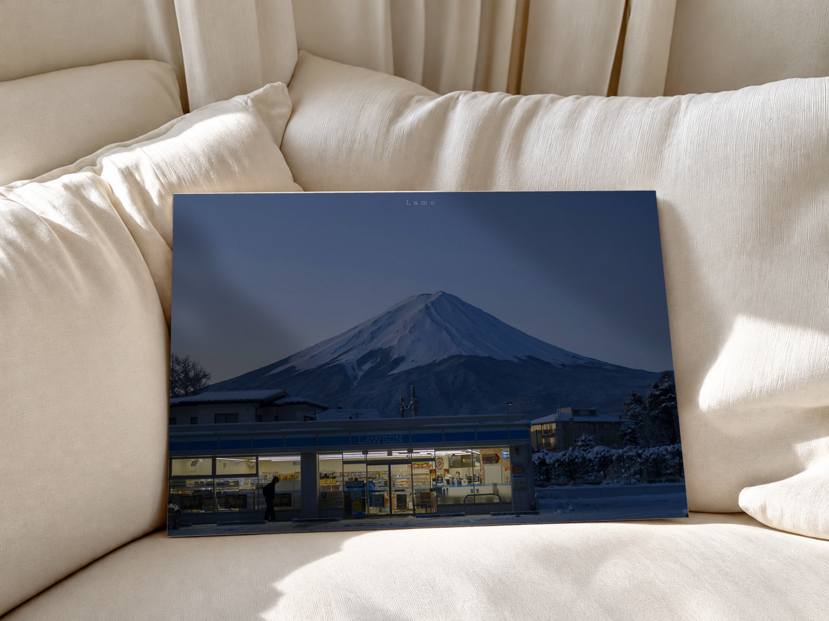 Warm beleuchteter Laden mit blauer Fassade unter einem verschneiten Berg im Abendblau, mit Person, Auto und Schnee. Ungerahmtes Poster an weiche weiße Sofakissen gelehnt.