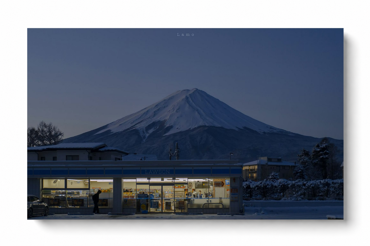 Warm beleuchteter Laden mit blauer Fassade unter einem verschneiten Berg im Abendblau, mit Person, Auto und Schnee. Ungerahmtes horizontales Poster vor weißem Hintergrund mit weichem Schatten.