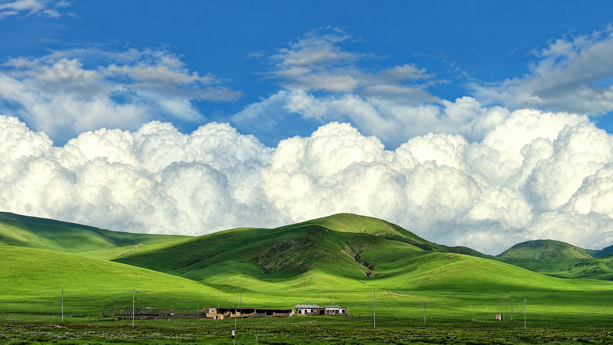 Colline verdi arrotondate e piccoli edifici rurali sotto grandi nuvole bianche e un cielo azzurro luminoso. Anteprima orizzontale senza cornice con la composizione completa.