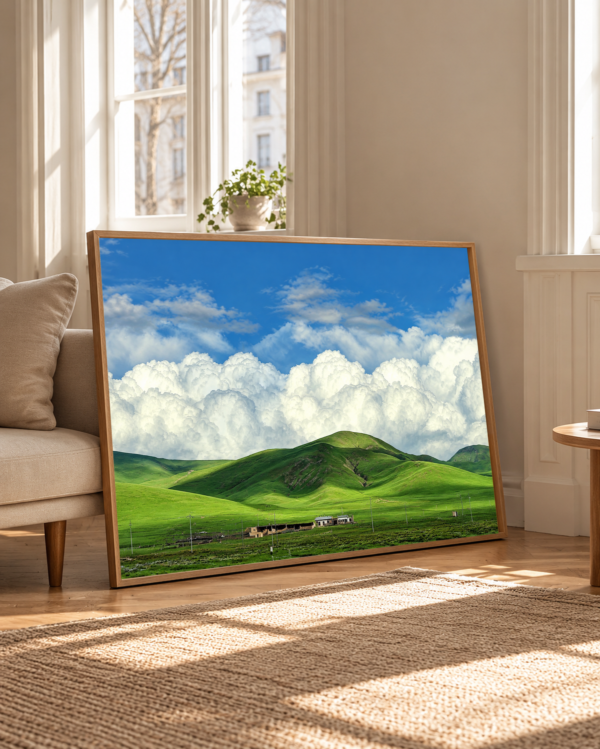 Colline verdi arrotondate e piccoli edifici rurali sotto grandi nuvole bianche e un cielo azzurro luminoso. Cornice dall’aspetto rovere accanto a una poltrona chiara e ad alte finestre nella calda luce naturale.