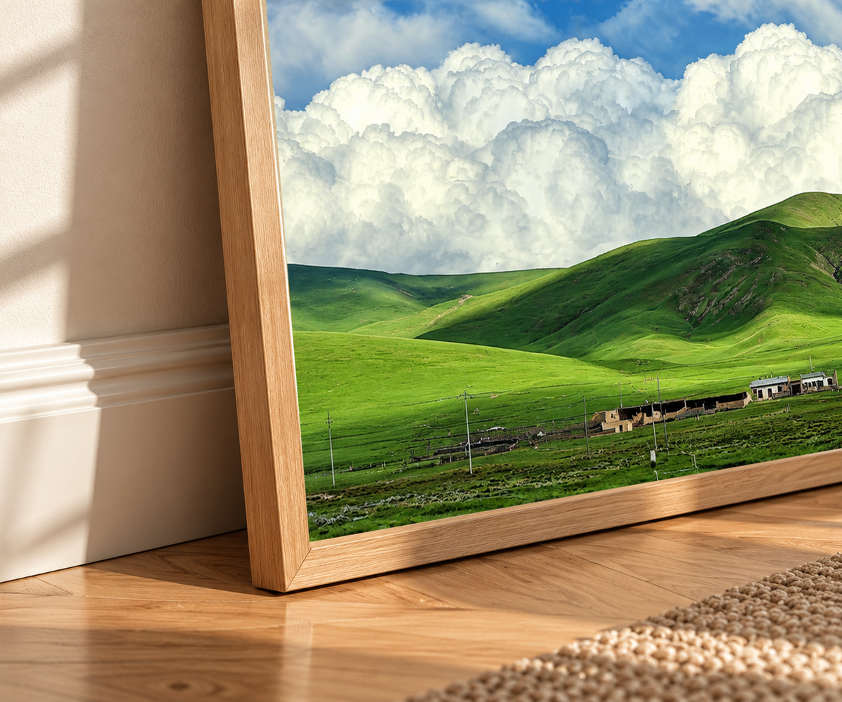 Colline verdi arrotondate e piccoli edifici rurali sotto grandi nuvole bianche e un cielo azzurro luminoso. Dettaglio di una cornice dall’aspetto rovere sul pavimento in legno, con venature della cornice visibili.
