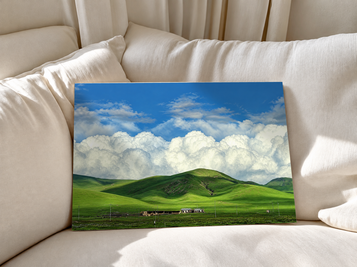 Colline verdi arrotondate e piccoli edifici rurali sotto grandi nuvole bianche e un cielo azzurro luminoso. Poster senza cornice appoggiato a morbidi cuscini bianchi del divano.