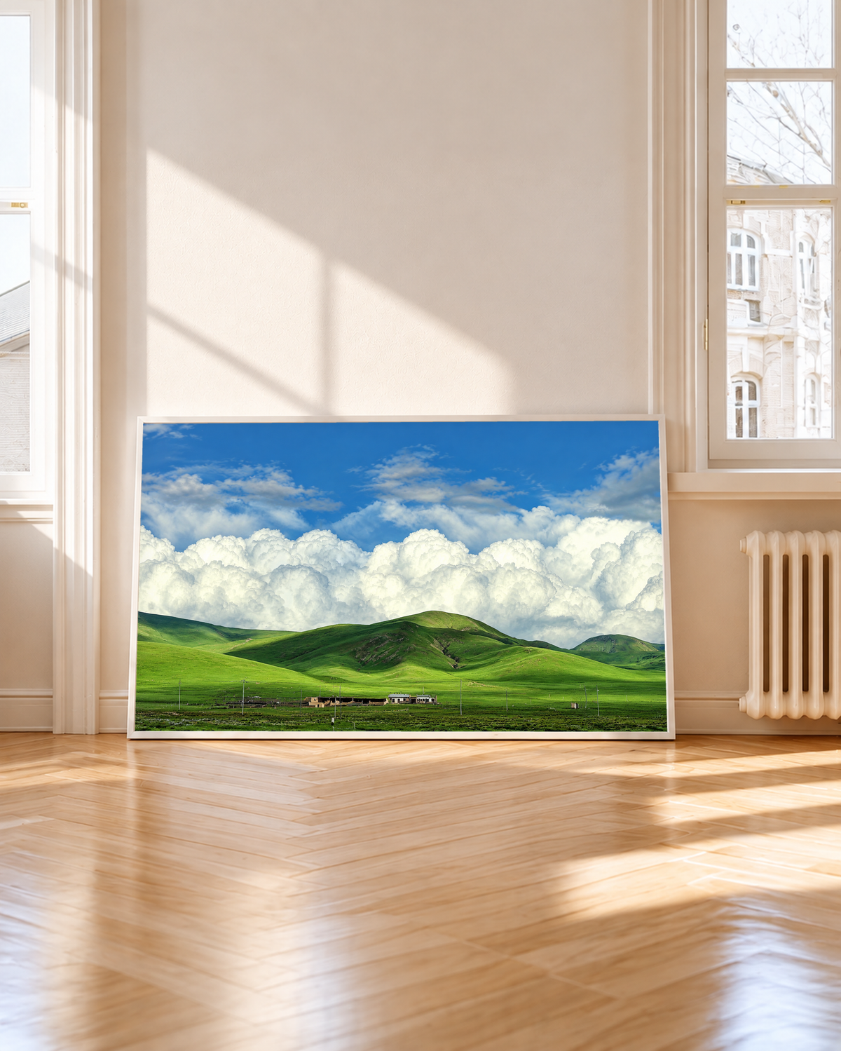 Colline verdi arrotondate e piccoli edifici rurali sotto grandi nuvole bianche e un cielo azzurro luminoso. Cornice bianca accanto a una finestra alta, con sole e ombre sul pavimento in legno.