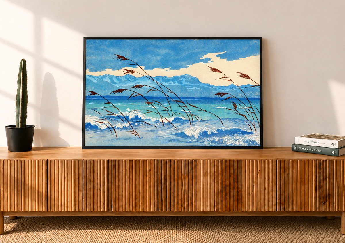 Russet reeds bend over curling blue and cream waves, with turquoise water, a distant mountain ridge and broad pale clouds behind. Black frame on a slatted wooden sideboard beside a potted cactus and books.