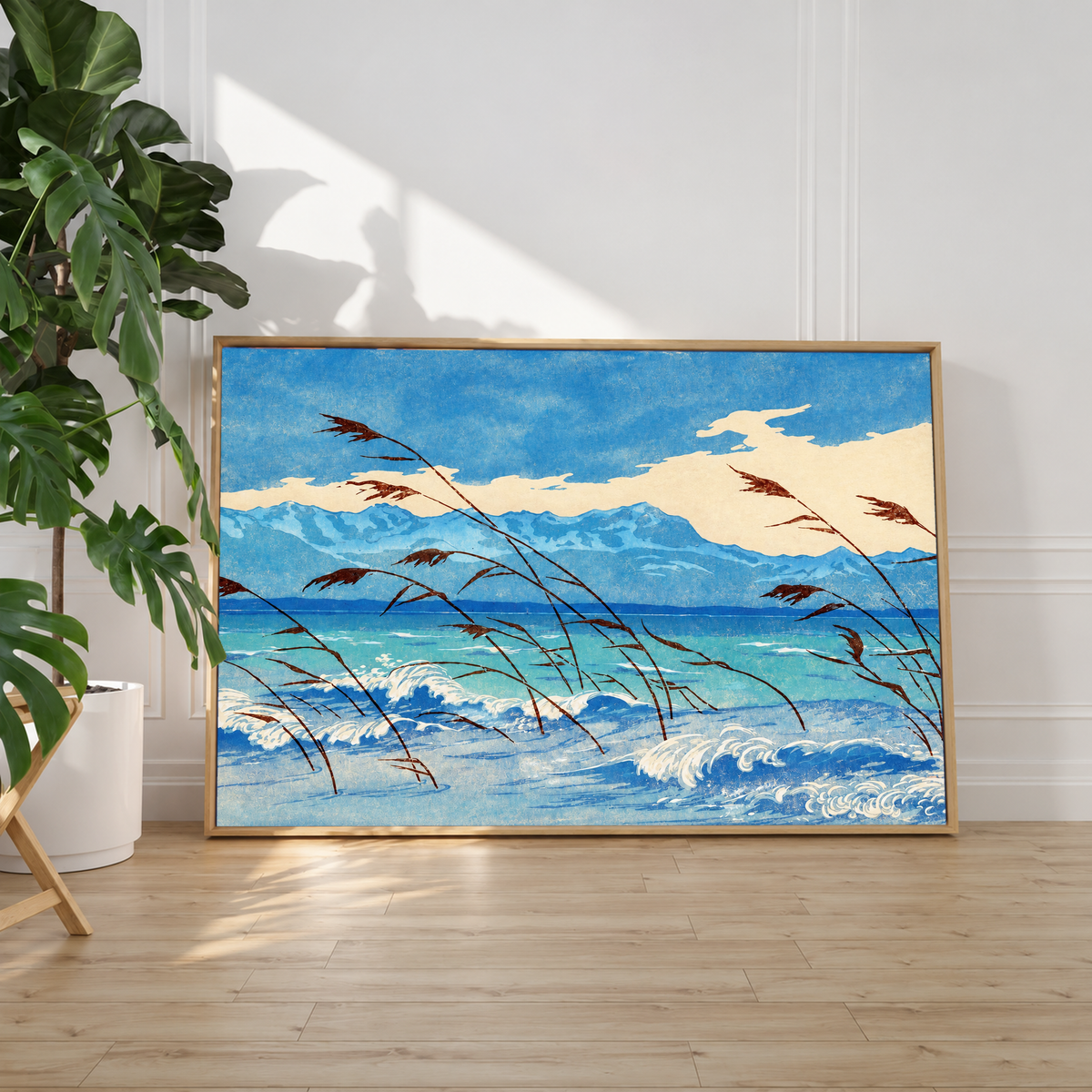 Russet reeds bend over curling blue and cream waves, with turquoise water, a distant mountain ridge and broad pale clouds behind. Light floating canvas frame leaning against a white wall beside a leafy houseplant, with sunlight on the wood floor.