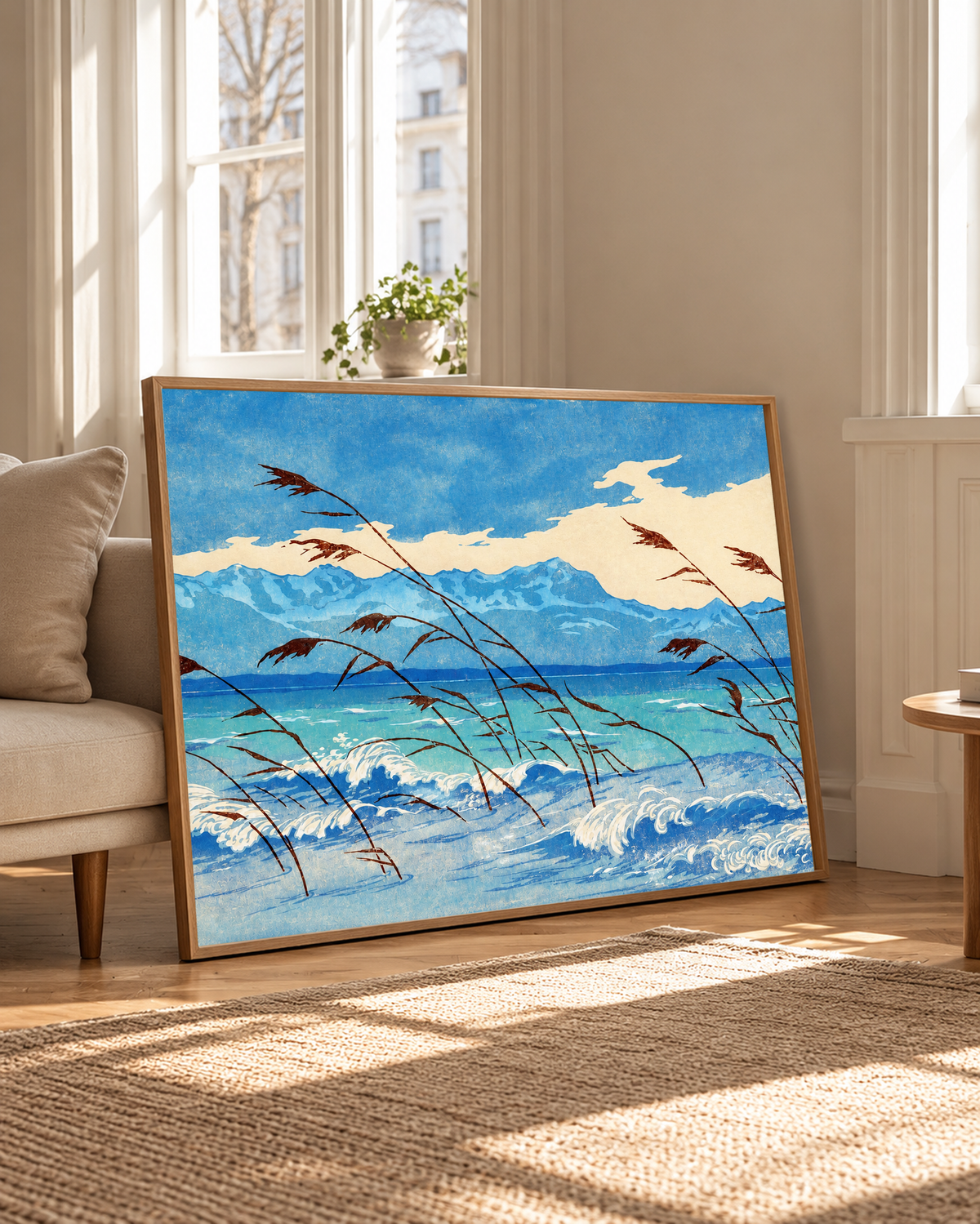 Russet reeds bend over curling blue and cream waves, with turquoise water, a distant mountain ridge and broad pale clouds behind. Oak frame beside a pale upholstered chair and tall windows in warm daylight.