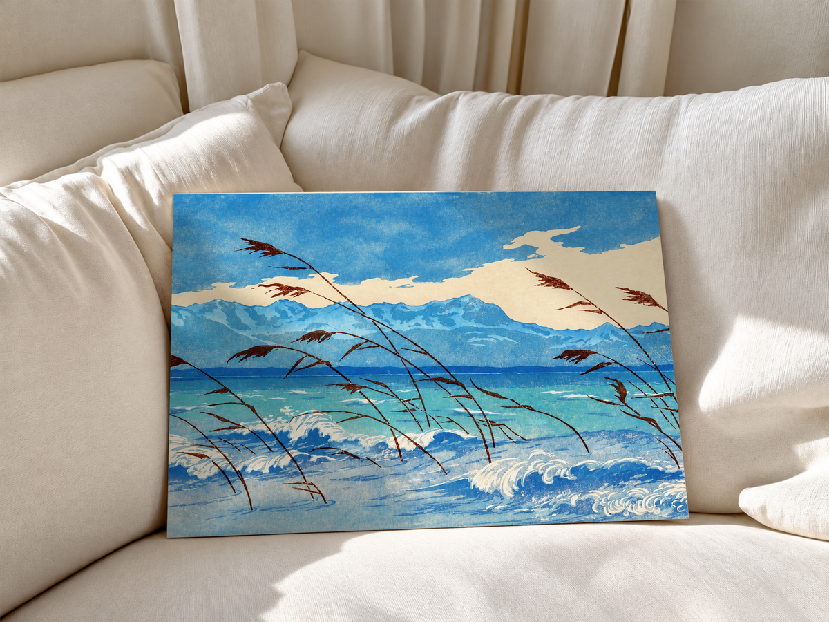 Russet reeds bend over curling blue and cream waves, with turquoise water, a distant mountain ridge and broad pale clouds behind. Unframed poster leaning against soft white sofa cushions.