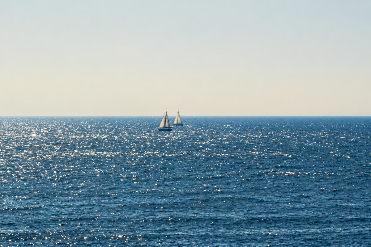 Due barche lontane dalle vele bianche vicino all’orizzonte sul mare blu scintillante sotto un ampio cielo chiaro crema-grigio. Anteprima orizzontale senza cornice con la composizione completa.