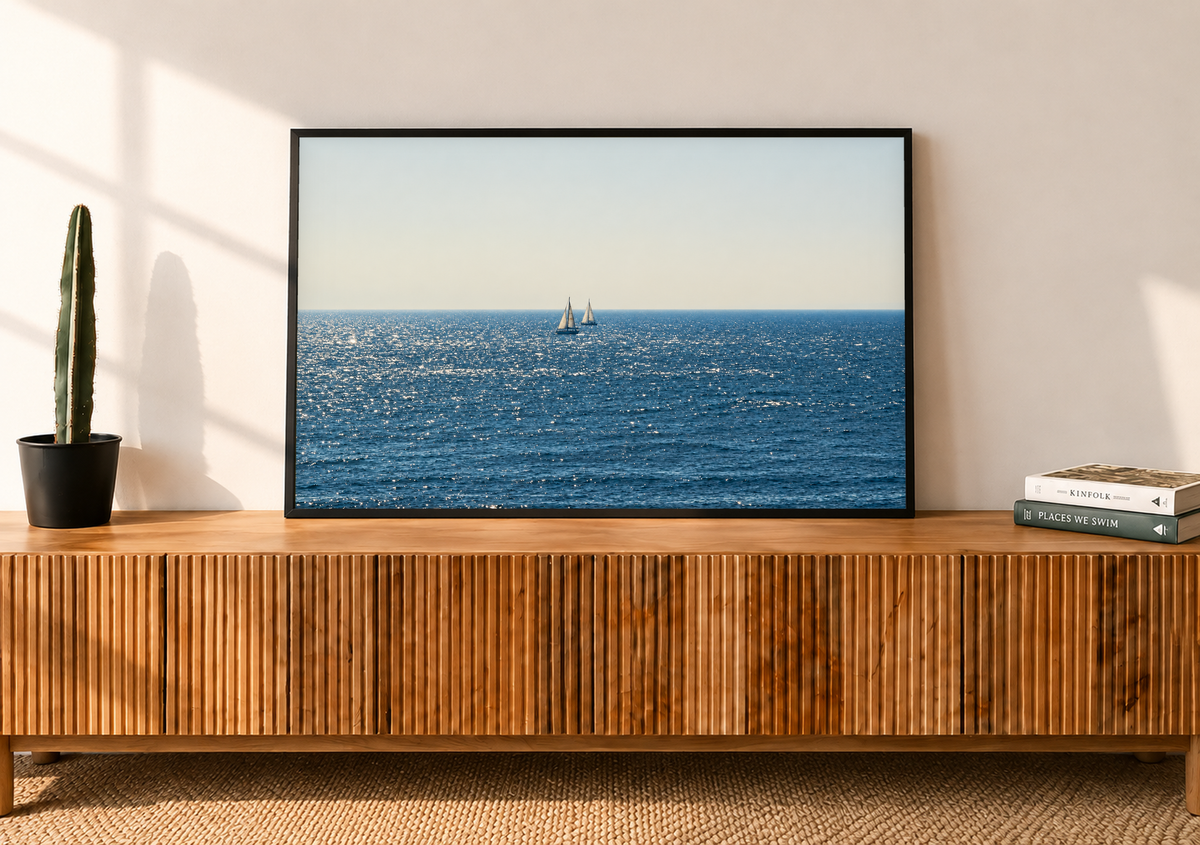 Due barche lontane dalle vele bianche vicino all’orizzonte sul mare blu scintillante sotto un ampio cielo chiaro crema-grigio. Cornice nera su una credenza in legno a listelli, accanto a un cactus in vaso e a libri.