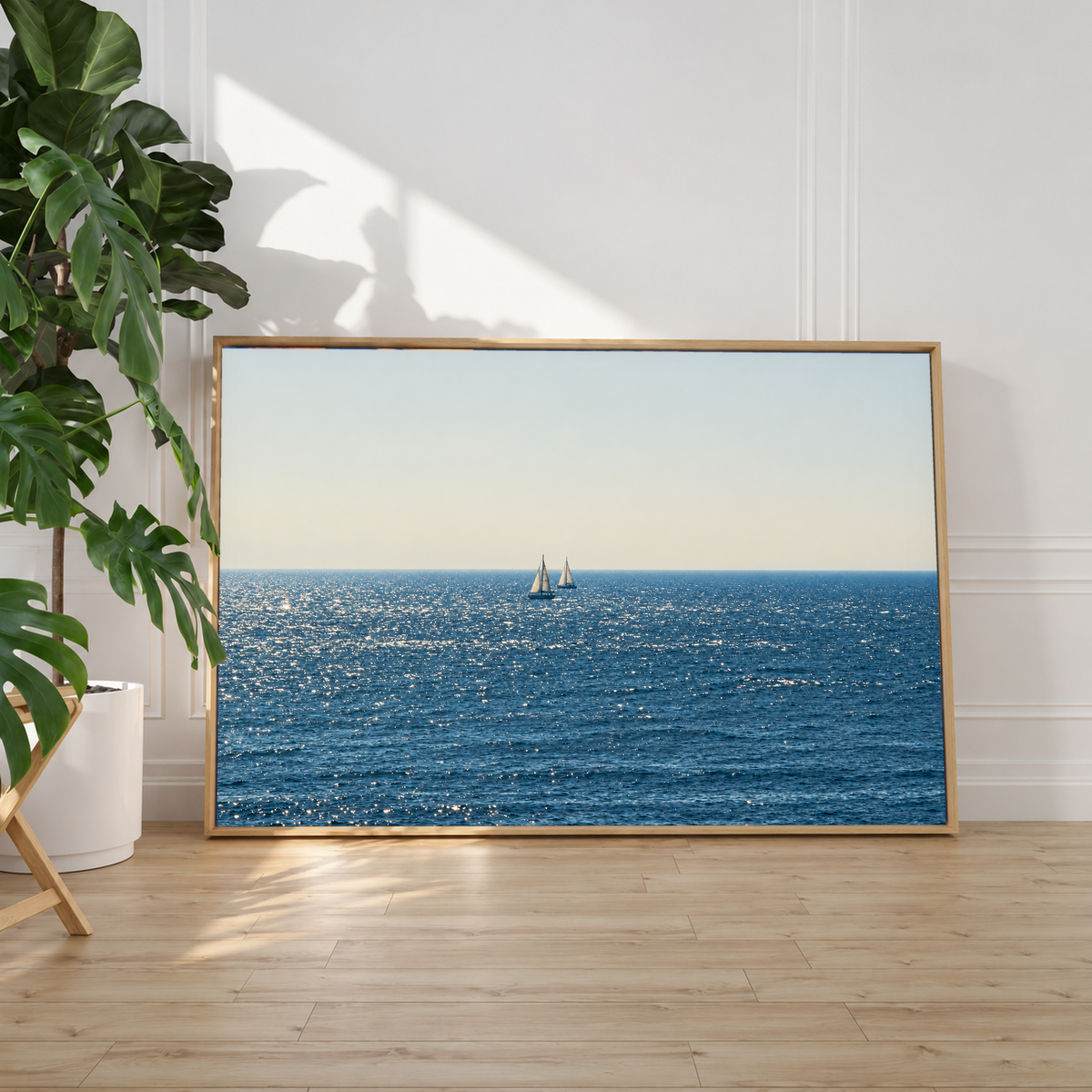 Due barche lontane dalle vele bianche vicino all’orizzonte sul mare blu scintillante sotto un ampio cielo chiaro crema-grigio. Cornice a cassetta chiara appoggiata a una parete bianca accanto a una pianta, con sole sul pavimento in legno.
