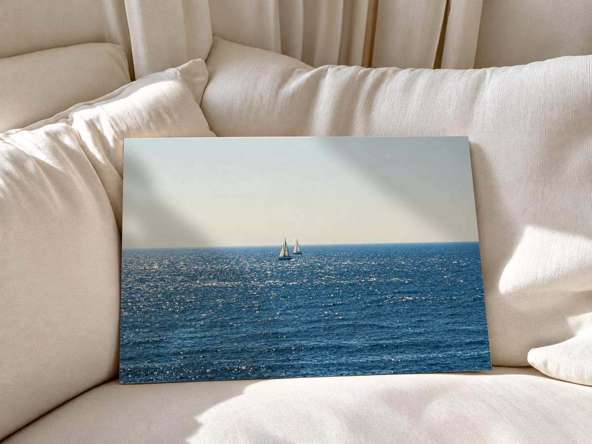 Due barche lontane dalle vele bianche vicino all’orizzonte sul mare blu scintillante sotto un ampio cielo chiaro crema-grigio. Poster senza cornice appoggiato a morbidi cuscini bianchi del divano.