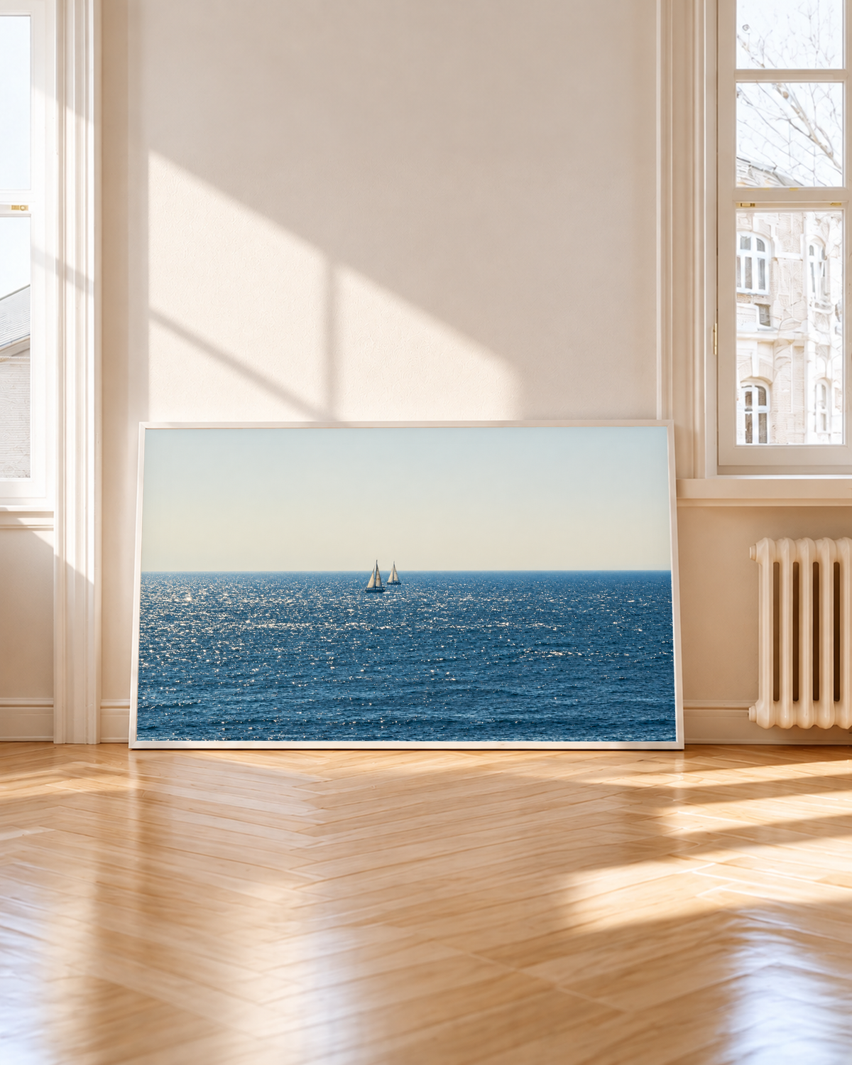 Due barche lontane dalle vele bianche vicino all’orizzonte sul mare blu scintillante sotto un ampio cielo chiaro crema-grigio. Cornice bianca accanto a una finestra alta, con sole e ombre sul pavimento in legno.