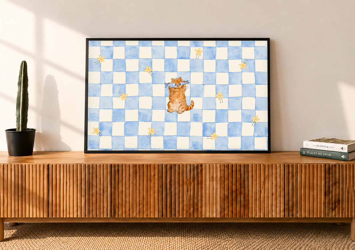 Un pequeño gato naranja con antifaz azul claro se sienta entre estrellas doradas sobre un tablero azul y crema. Marco negro sobre un aparador de madera con listones, junto a un cactus en maceta y libros.